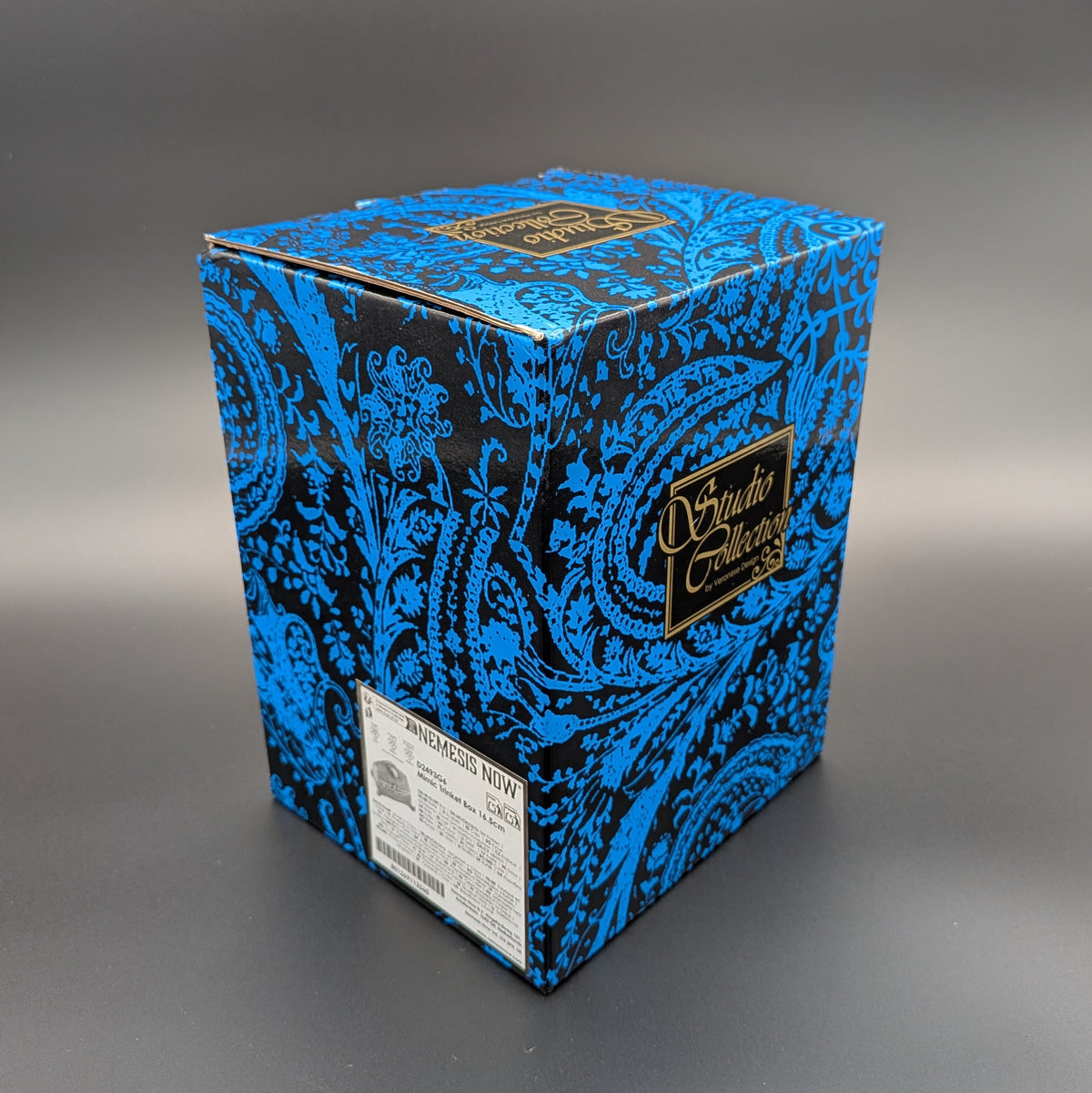 A square cardboard product box with a vibrant blue and black paisley pattern, featuring a gold 'Sudio Collection' logo on the side and a white 'NEMESIS NOW' label on the bottom edge, set against a dark gray gradient background.