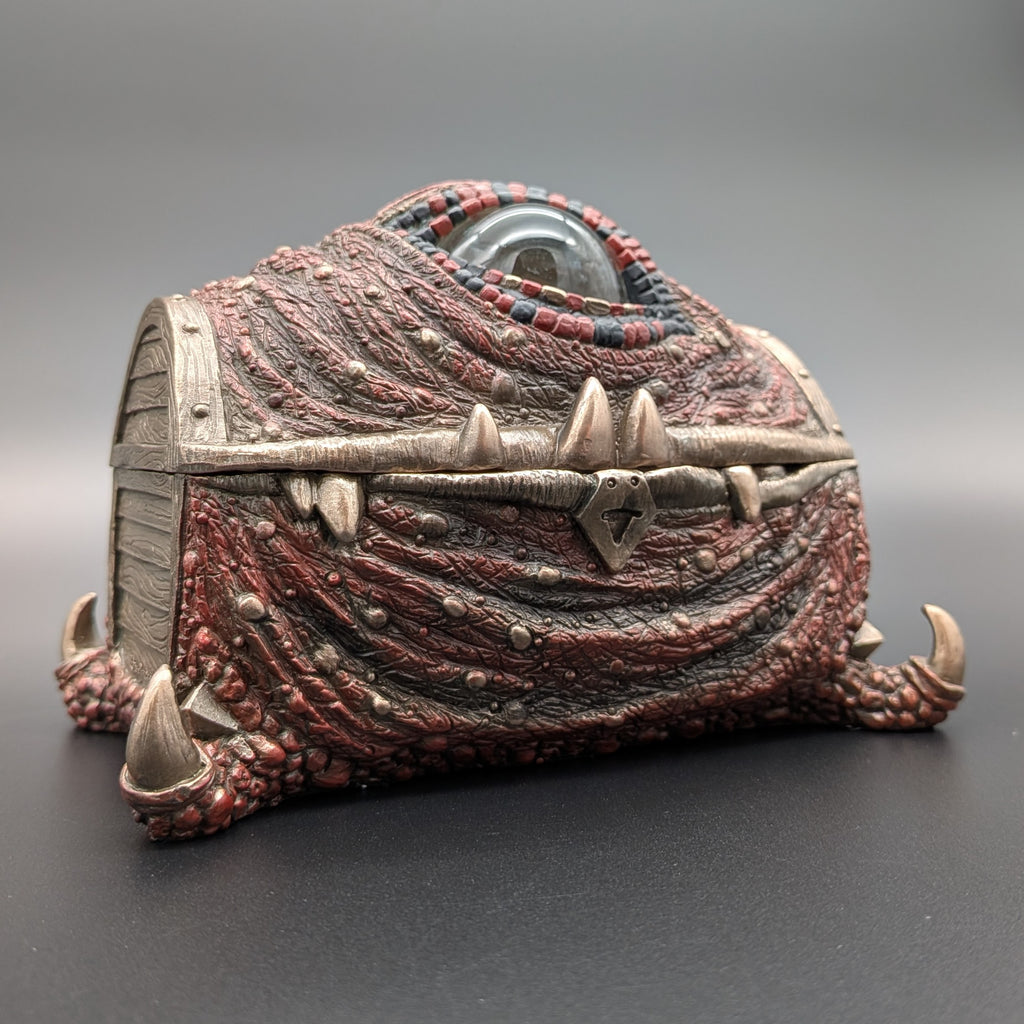 A fantasy monster eye chest figurine with textured red and dark gray scales, claws and a single large eye on top, shown on a dark gray background.