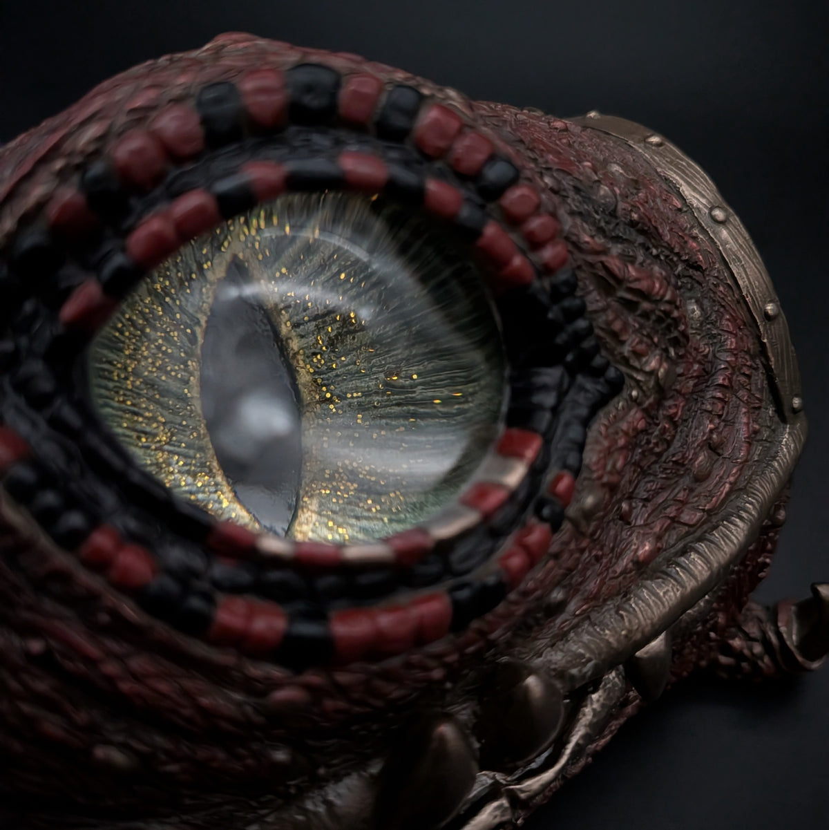 A close-up of a highly detailed fantasy eye accessory, featuring a realistic reptilian eye with a gold glittery iris and a dark pupil, surrounded by a textured reddish-brown and black frame with metallic bronze accents.