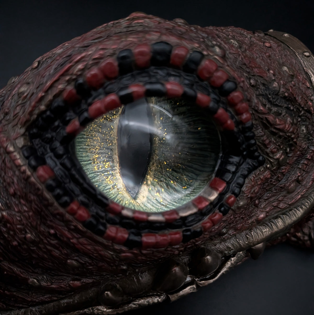 Close-up of a highly detailed fantasy creature eye with a large slit pupil, shimmering gold and green iris, surrounded by a black and red beaded ring, set against a dark textured reptilian head.