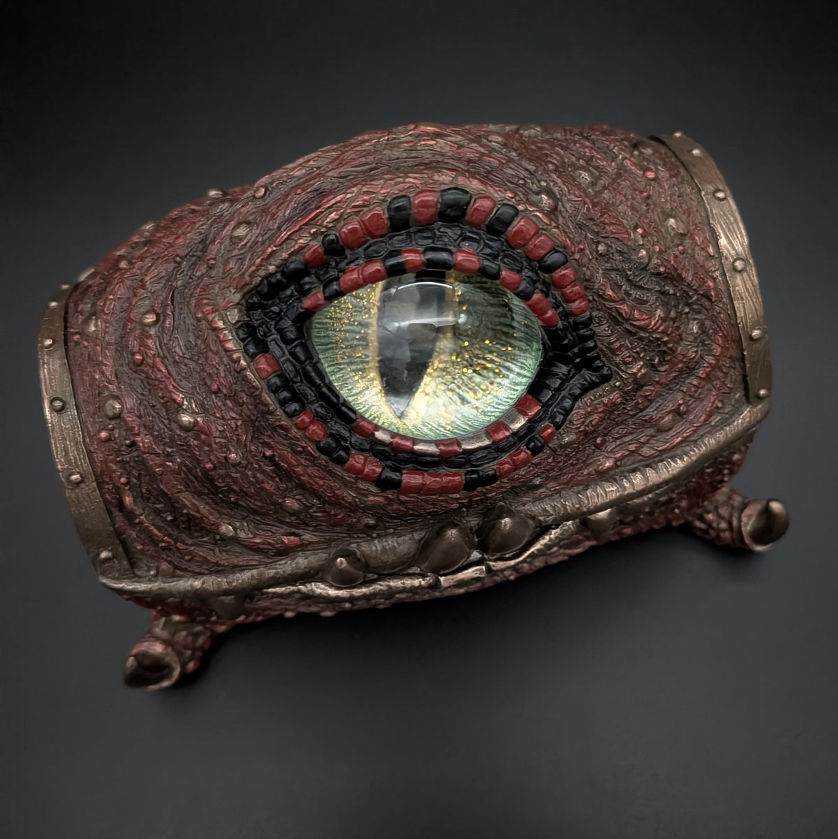 A highly detailed fantasy collectible figurine of a mimic chest, featuring a textured reddish-brown scaly head, metallic bronze bands, and a large green and gold iris surrounded by red and black scales, set against a dark background.