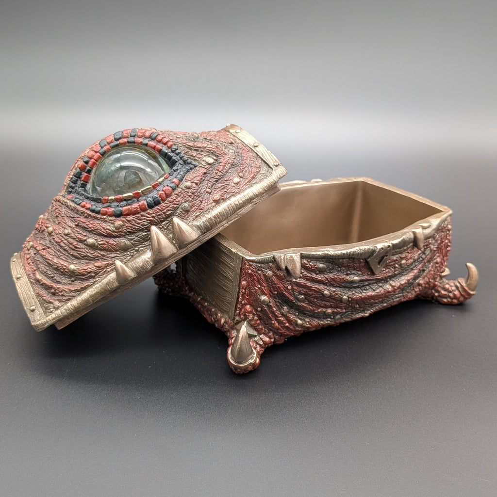 A fantasy creature-eye jewelry box with a textured reddish-brown and bronze body, featuring a glass eye on the lid, set against a dark gray background.