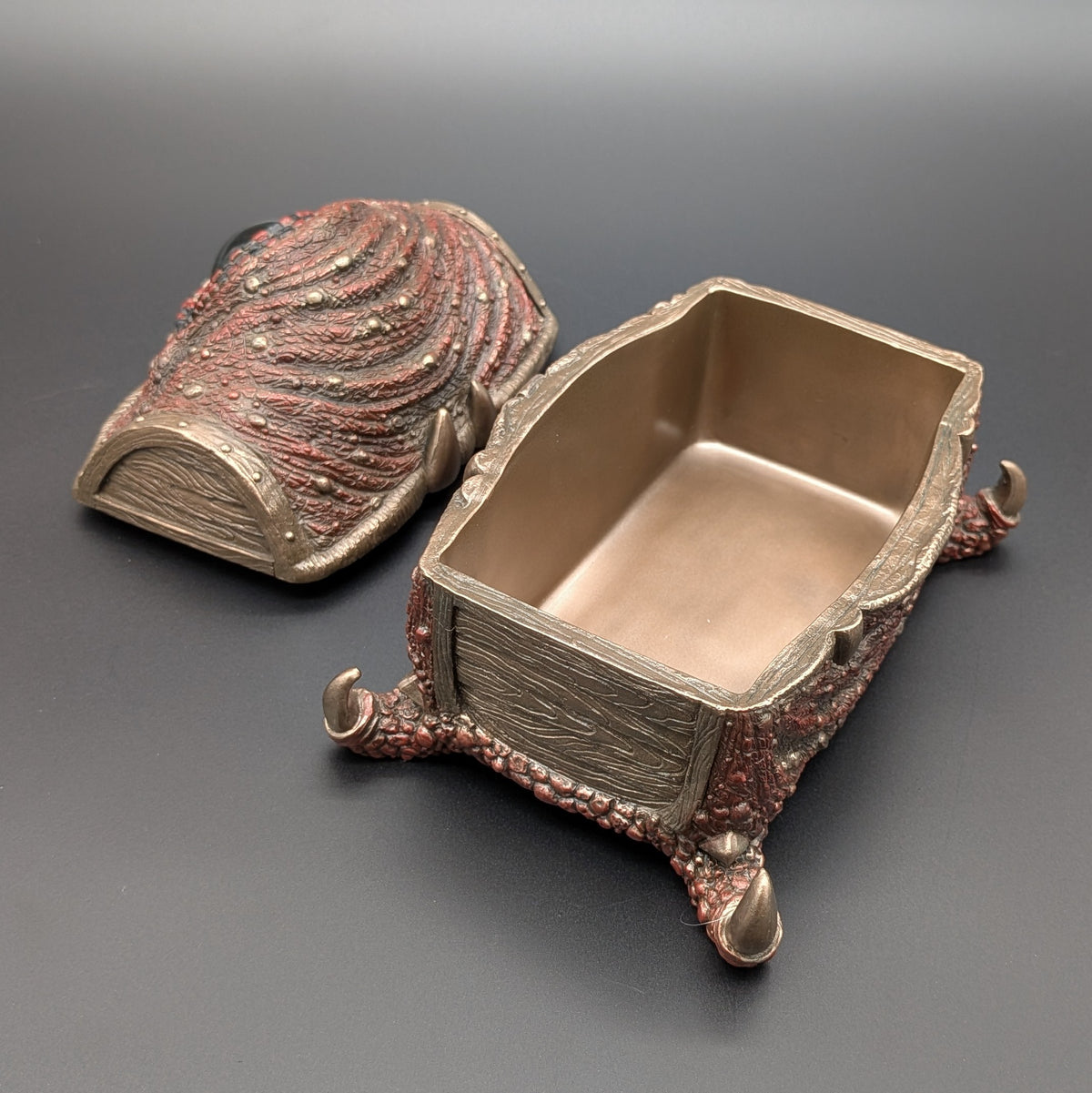 An ornate box with a textured, reptile-like surface in silver and copper tones, shown open with its lid placed beside it on a dark gray background.