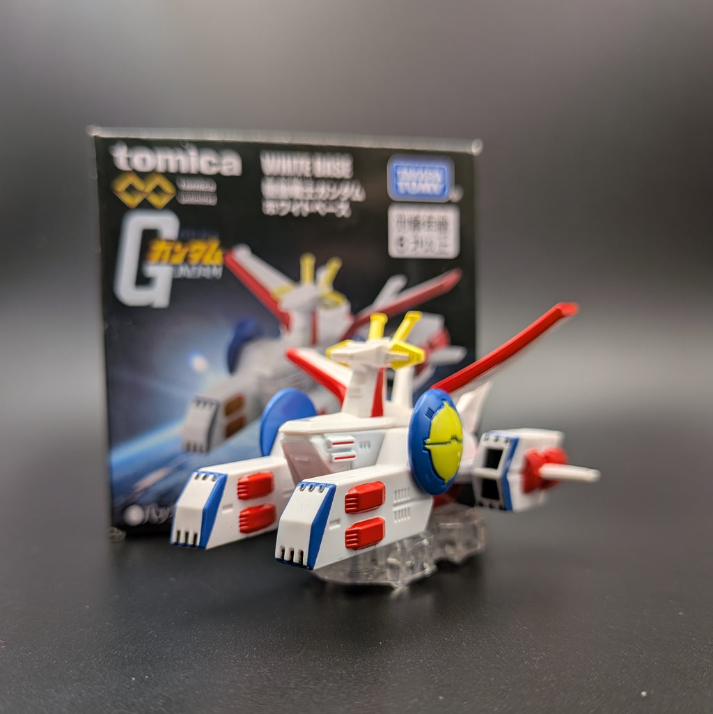 A Tomica Mobile Suit Gundam White Base toy model displayed in front of its original packaging box on a dark gray surface.