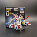A Tomica Mobile Suit Gundam White Base toy model displayed in front of its original packaging box on a dark gray surface.