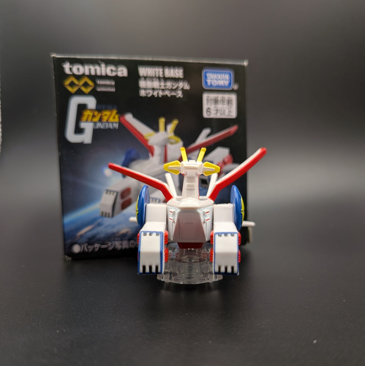 A small Tomica Gundam model toy in front of its retail box on a dark surface. The box reads Tomica and Gundam White Base, and the toy itself is primarily white with red, blue, and yellow accents.