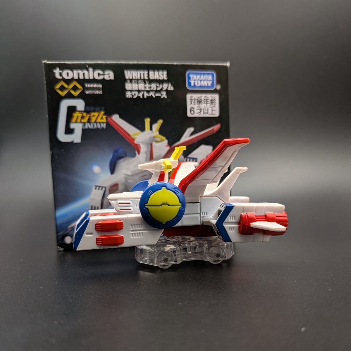 TOMICA Gundam White Base toy model with its original packaging box, displayed on a dark gray surface.