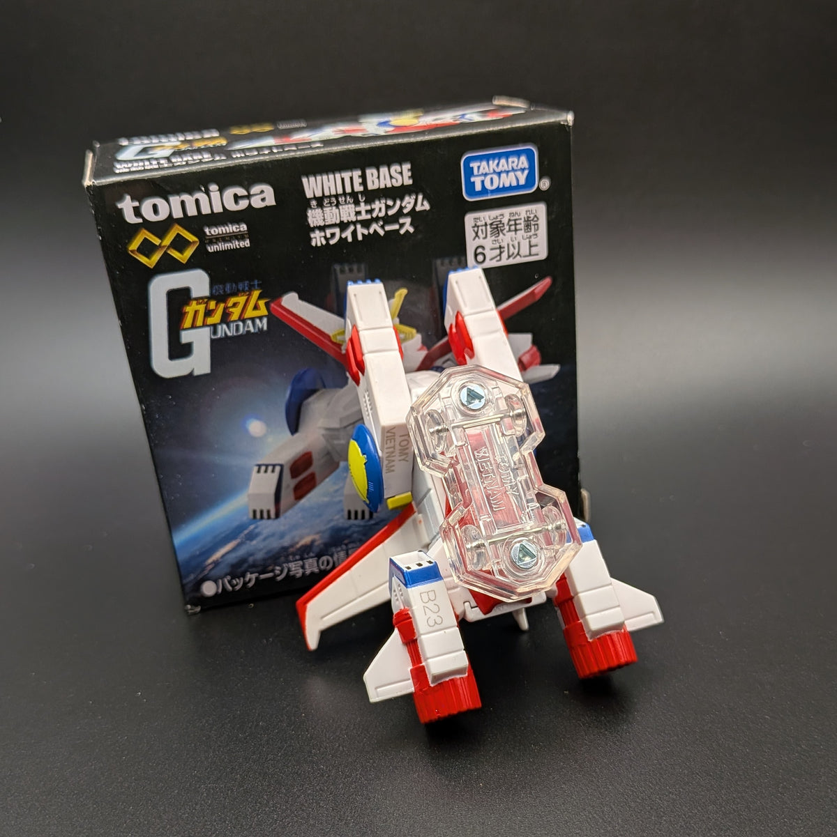 Tomica Gundam White Base toy model shown in front of its retail box on a dark surface. The model is white with red and blue accents and has a clear plastic base.