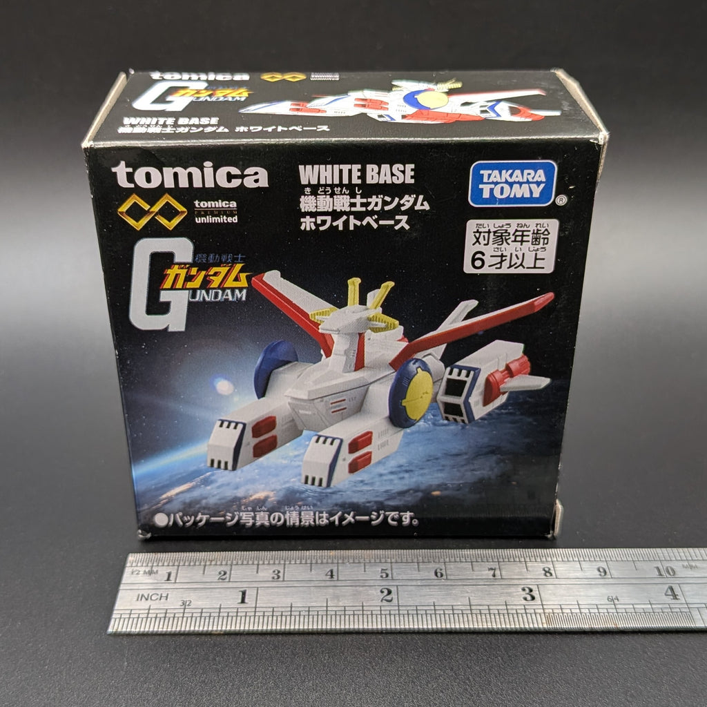 Boxed TOMICA Mobile Suit Gundam White Base toy box by Takara Tomy, shown upright on a dark surface with a metal ruler in front for scale. The packaging features a printed model vehicle and visible TOMICA and Takara Tomy branding.