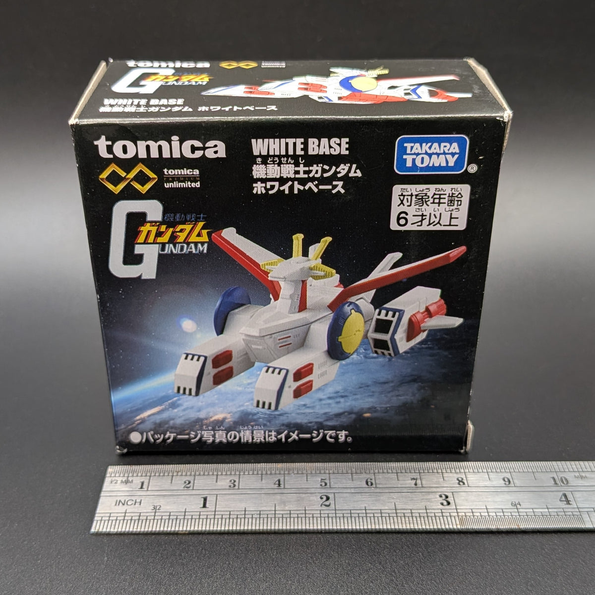 Boxed TOMICA Mobile Suit Gundam White Base toy box by Takara Tomy, shown upright on a dark surface with a metal ruler in front for scale. The packaging features a printed model vehicle and visible TOMICA and Takara Tomy branding.