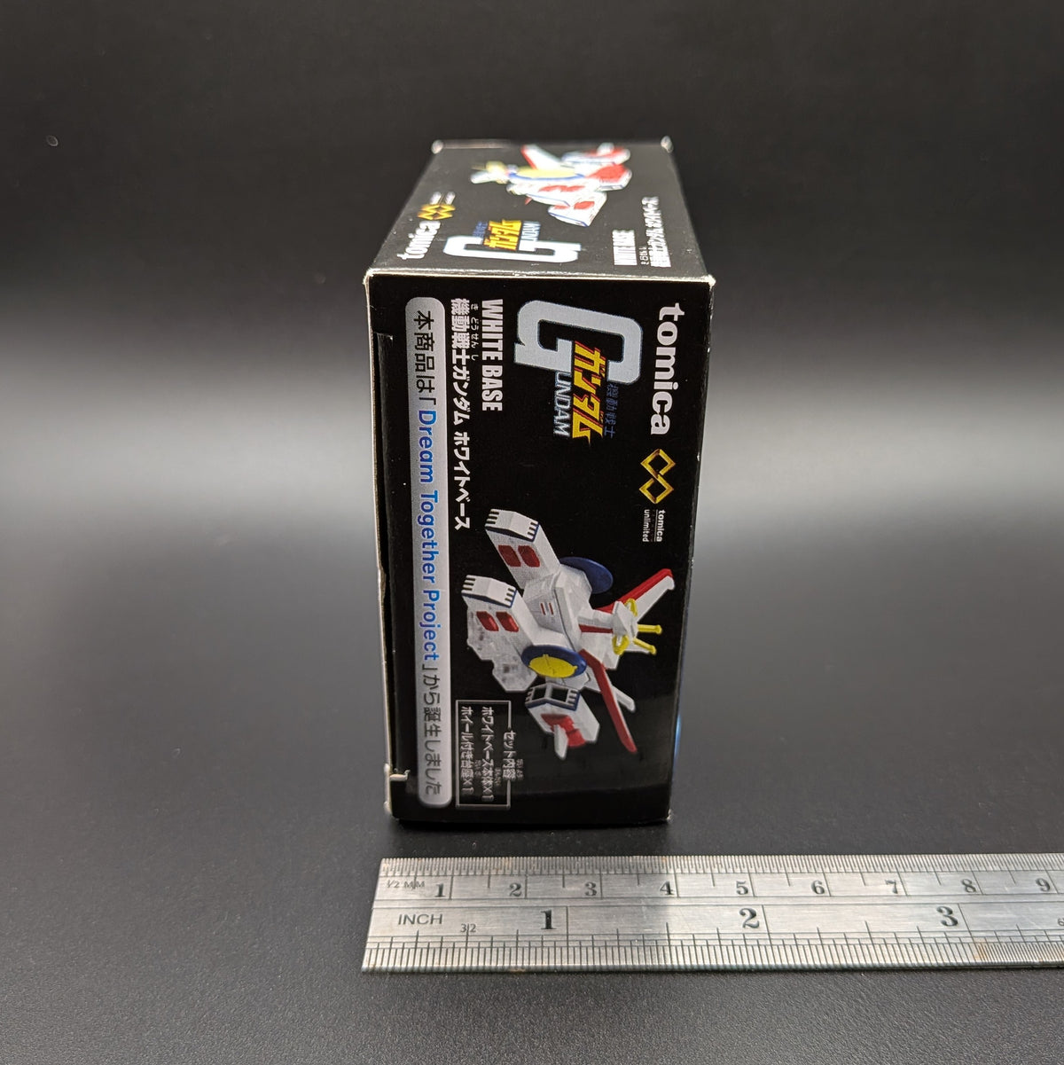 A boxed toy model stands upright on a dark surface with a metal ruler below it for scale. The box is predominantly black with white, red, and yellow printed graphics and text, including the visible brand name Tomica.