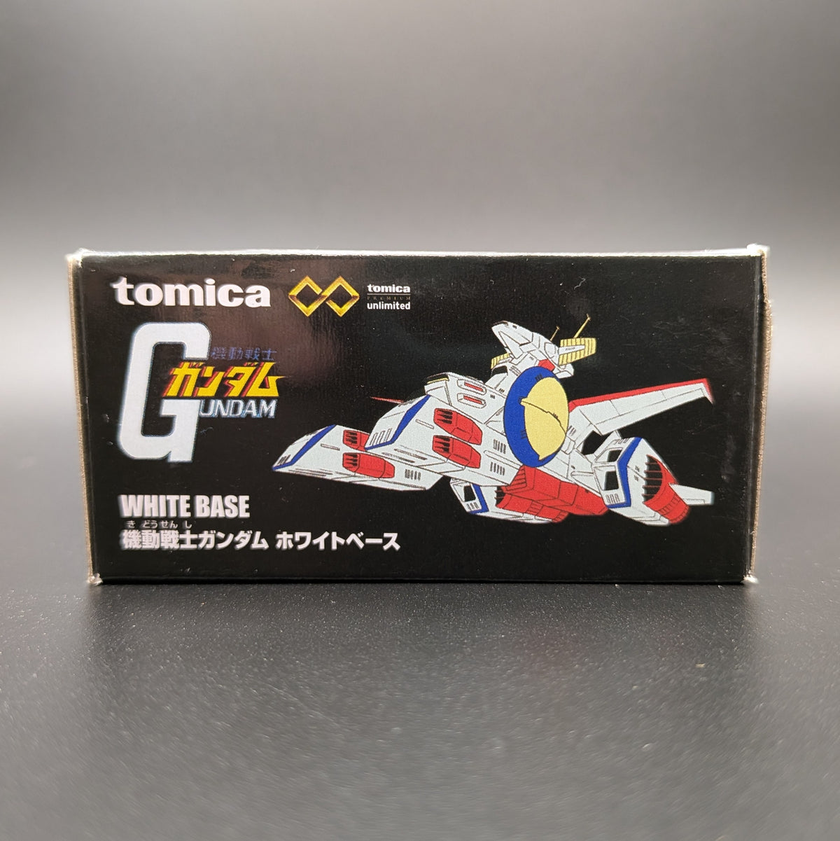 Tomica Gundam White Base  packaging box, featuring a black background with white, red, and blue Gundam illustrations and Japanese text.