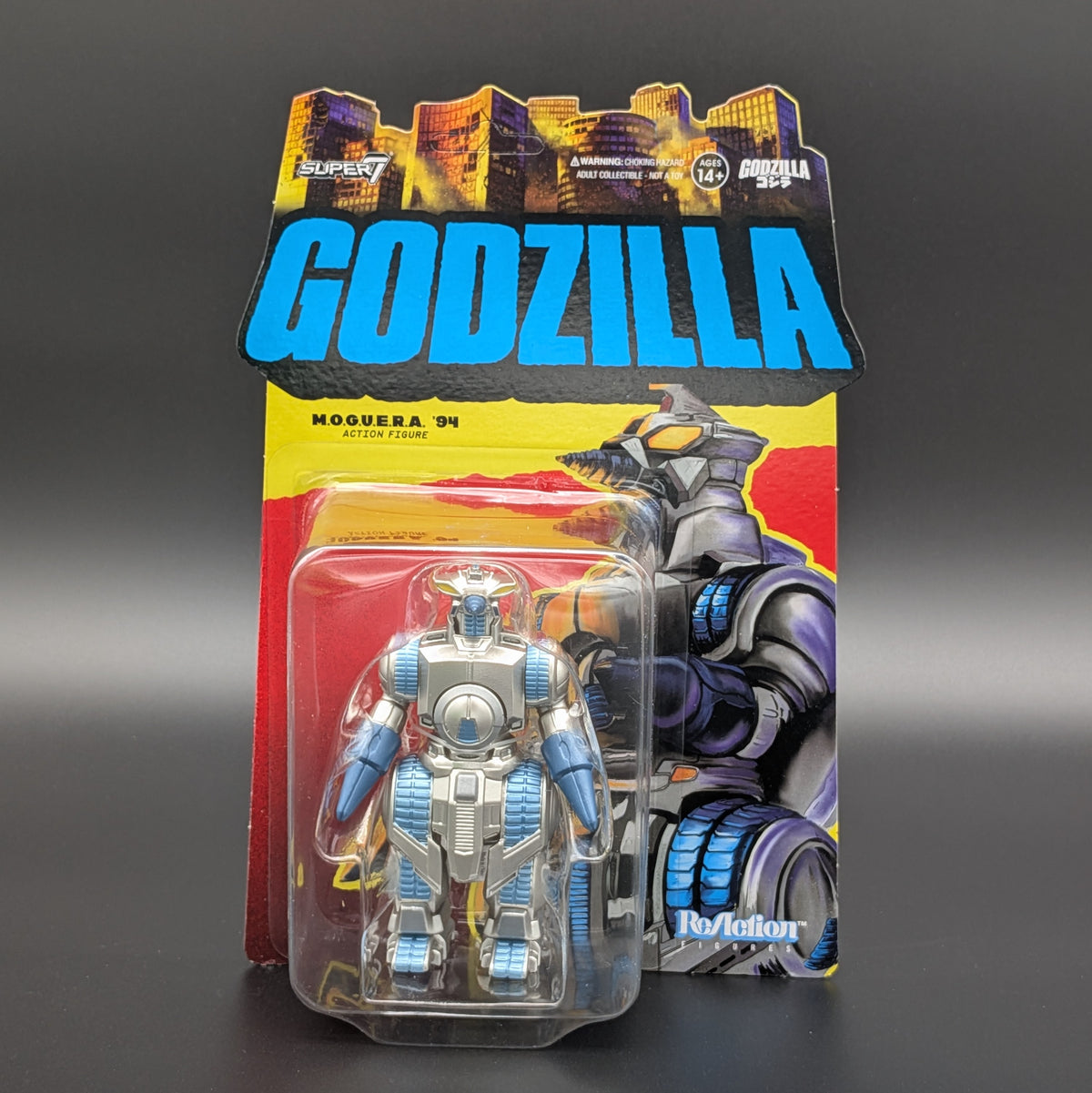 A packaged Godzilla action figure set on a dark gray background. The cardback features a large blue GODZILLA logo, Godzilla artwork, and a clear blister pack holding a silver and blue robot figure.