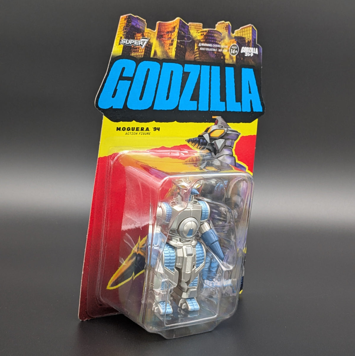 A packaged Godzilla Moguera '94 action figure in a clear blister pack with a colorful cardback. The cardback features a large blue 'GODZILLA' logo, a yellow band, and red lower sections, with the figure visible inside.