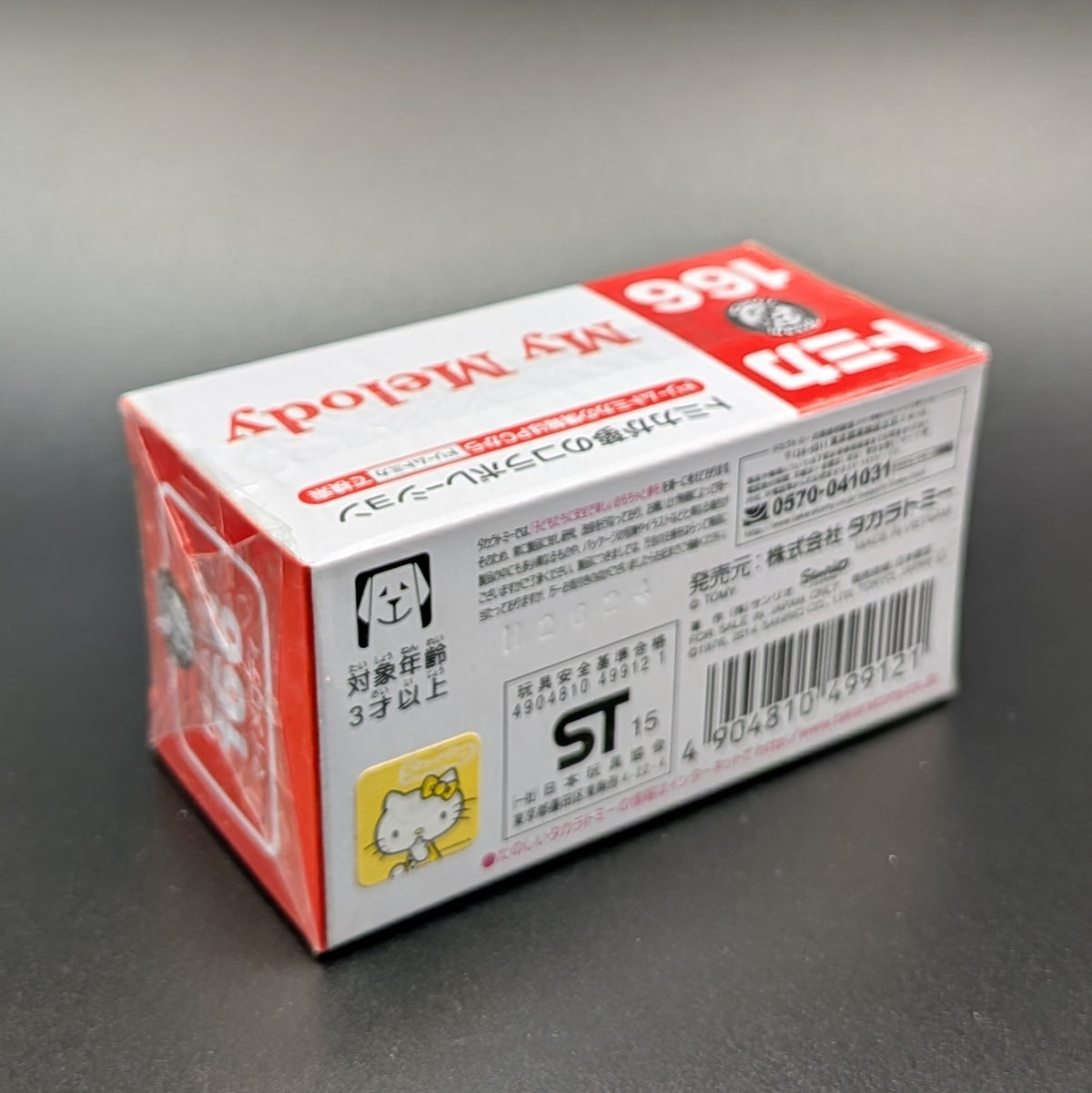 A boxed package photographed on a dark gray surface. The box is predominantly white with red panels and black text, featuring Hello Kitty branding and Japanese text.