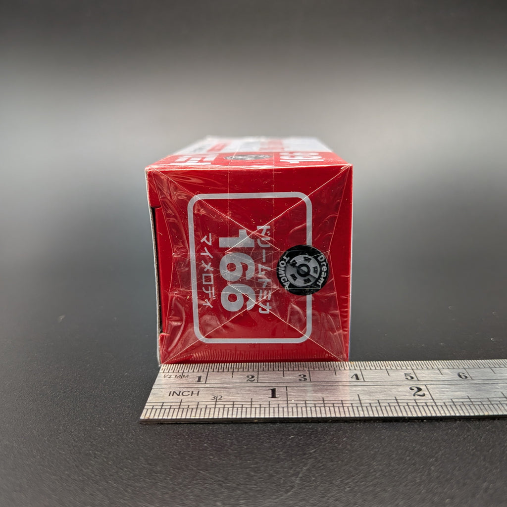 A small red product box wrapped in clear plastic, standing upright on a dark surface with a metal ruler underneath for scale. The box has white Japanese text and a black circular logo, on a gray gradient background.