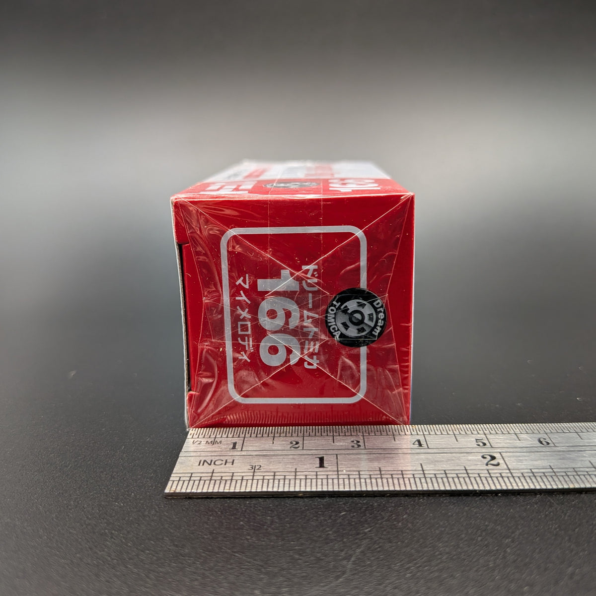 A small red product box wrapped in clear plastic, standing upright on a dark surface with a metal ruler underneath for scale. The box has white Japanese text and a black circular logo, on a gray gradient background.