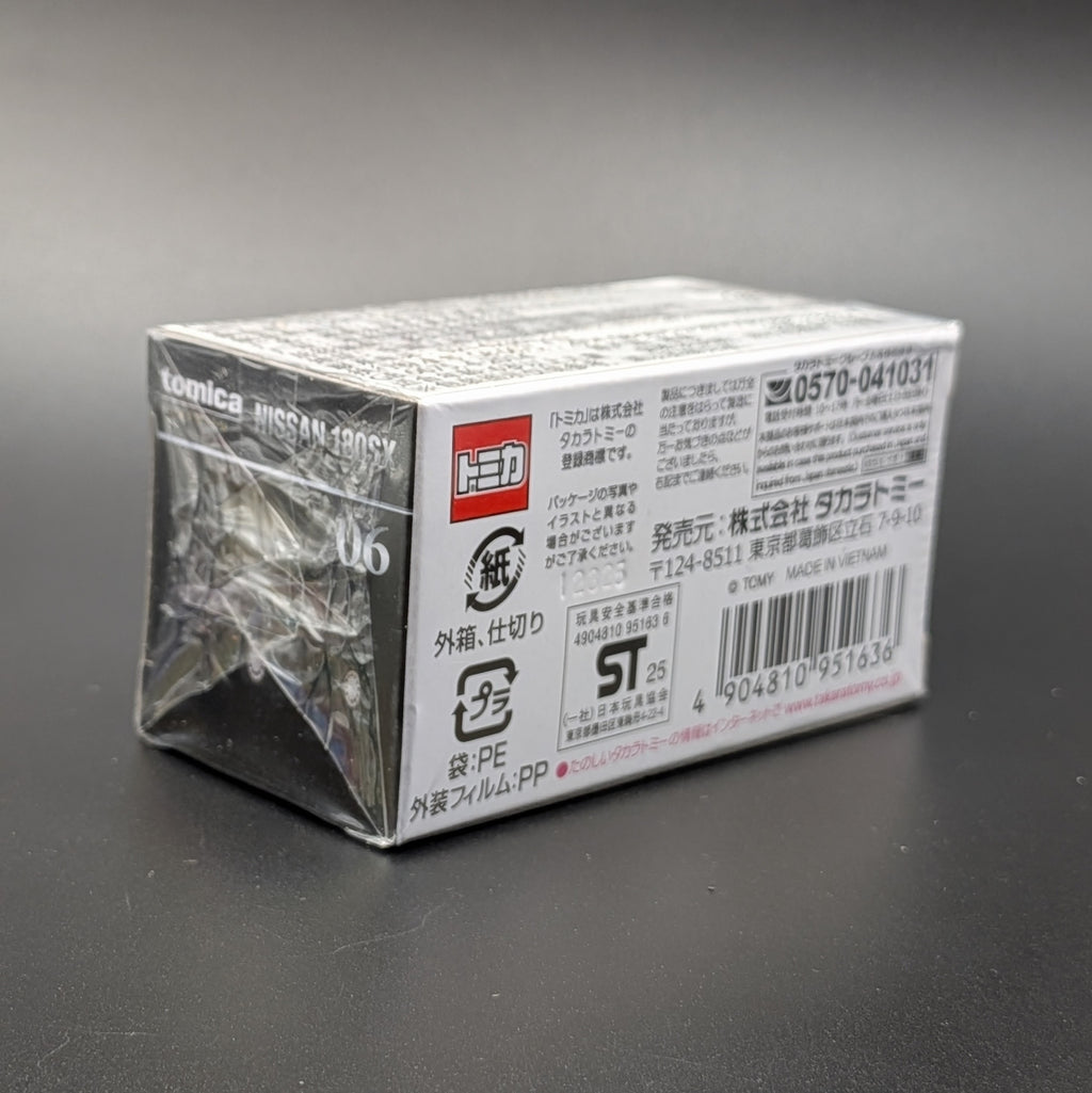 A boxed toy product wrapped in clear plastic, photographed on a dark gray surface. The package has printed text and a red Tomica logo on the side.