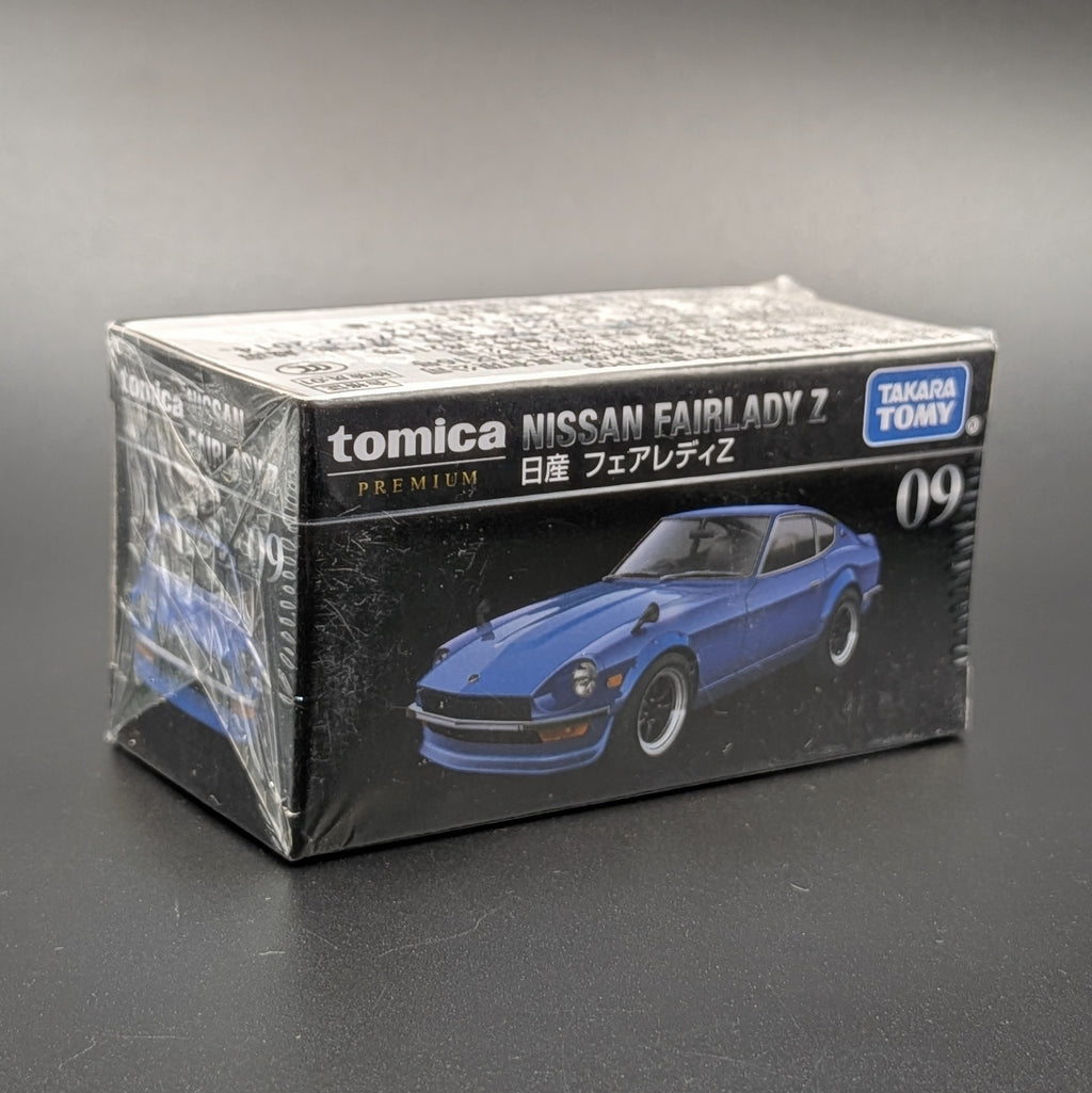 A boxed Tomica Nissan Fairlady Z model car package, sealed in clear plastic, photographed on a dark gray surface against a gray background. The box is black with white text and shows a blue car illustration, with visible brands Tomica and Takara Toy.