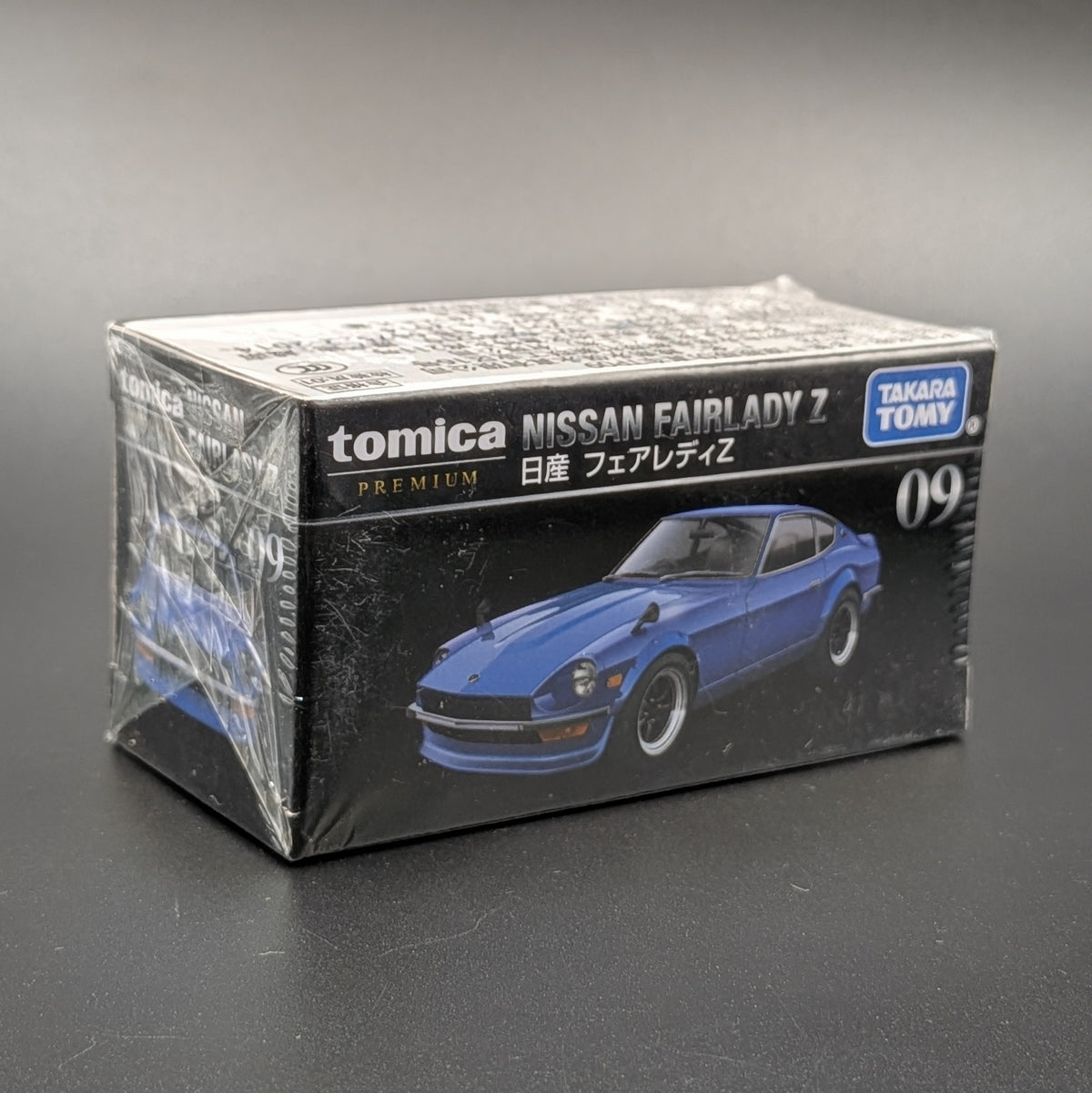 A boxed Tomica Nissan Fairlady Z model car package, sealed in clear plastic, photographed on a dark gray surface against a gray background. The box is black with white text and shows a blue car illustration, with visible brands Tomica and Takara Toy.