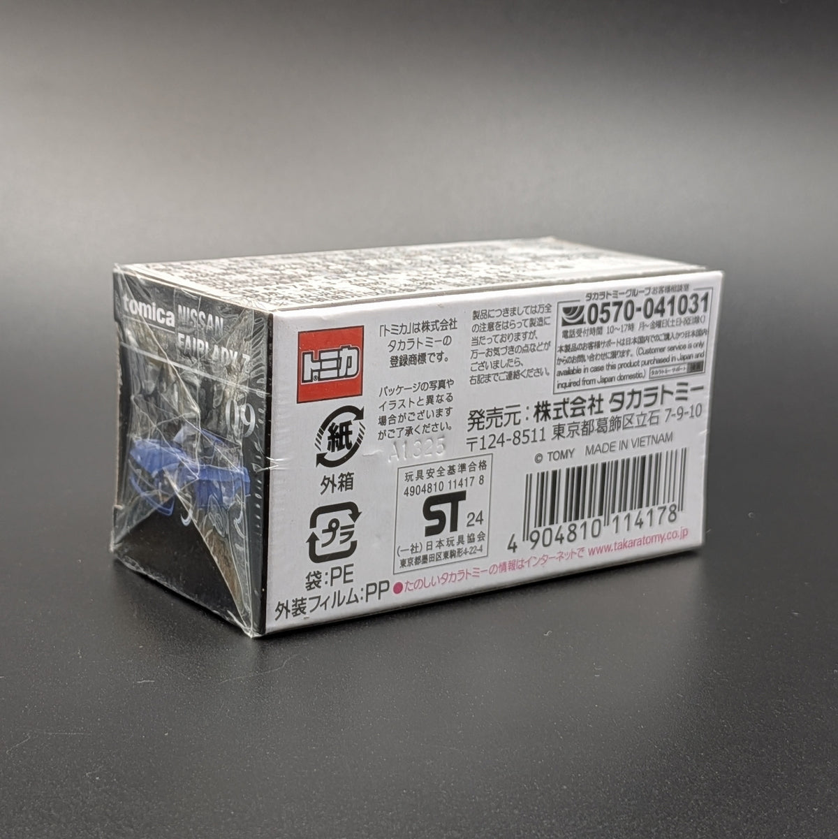 A sealed plastic-wrapped box of a Tomica scale model, photographed on a dark gray background.