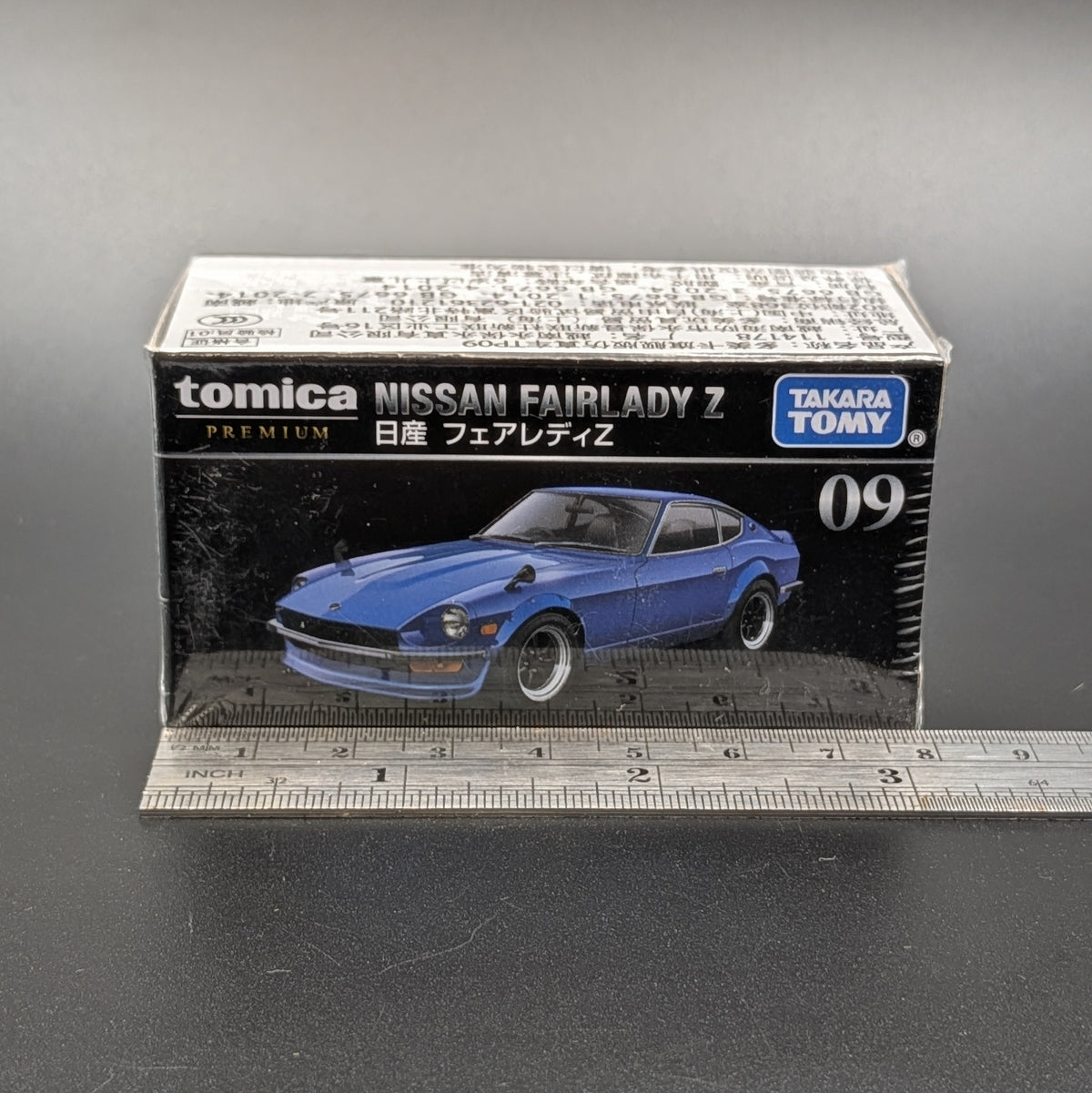 A boxed Tomica Premium Nissan Fairlady Z model car, shown in its retail packaging with a ruler in front for scale on a dark gray surface.