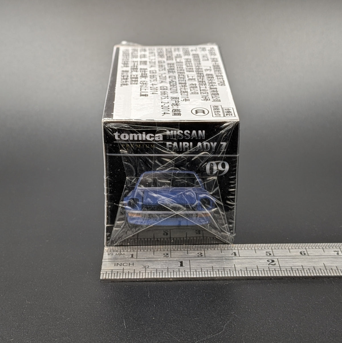 A boxed Tomica Nissan Fairlady 70 die-cast model car in its original packaging, wrapped in protective plastic, with a metal ruler placed underneath to show scale on a dark gray background.