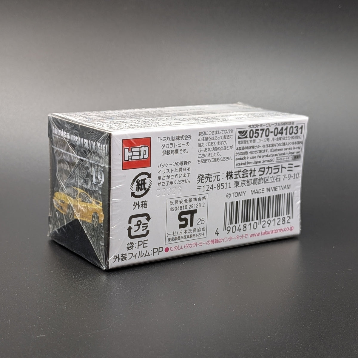 A boxed toy product wrapped in clear plastic, photographed on a dark gray background. The package is mostly white with black text, a red logo block, and a barcode, and a small yellow toy is visible on the left side.