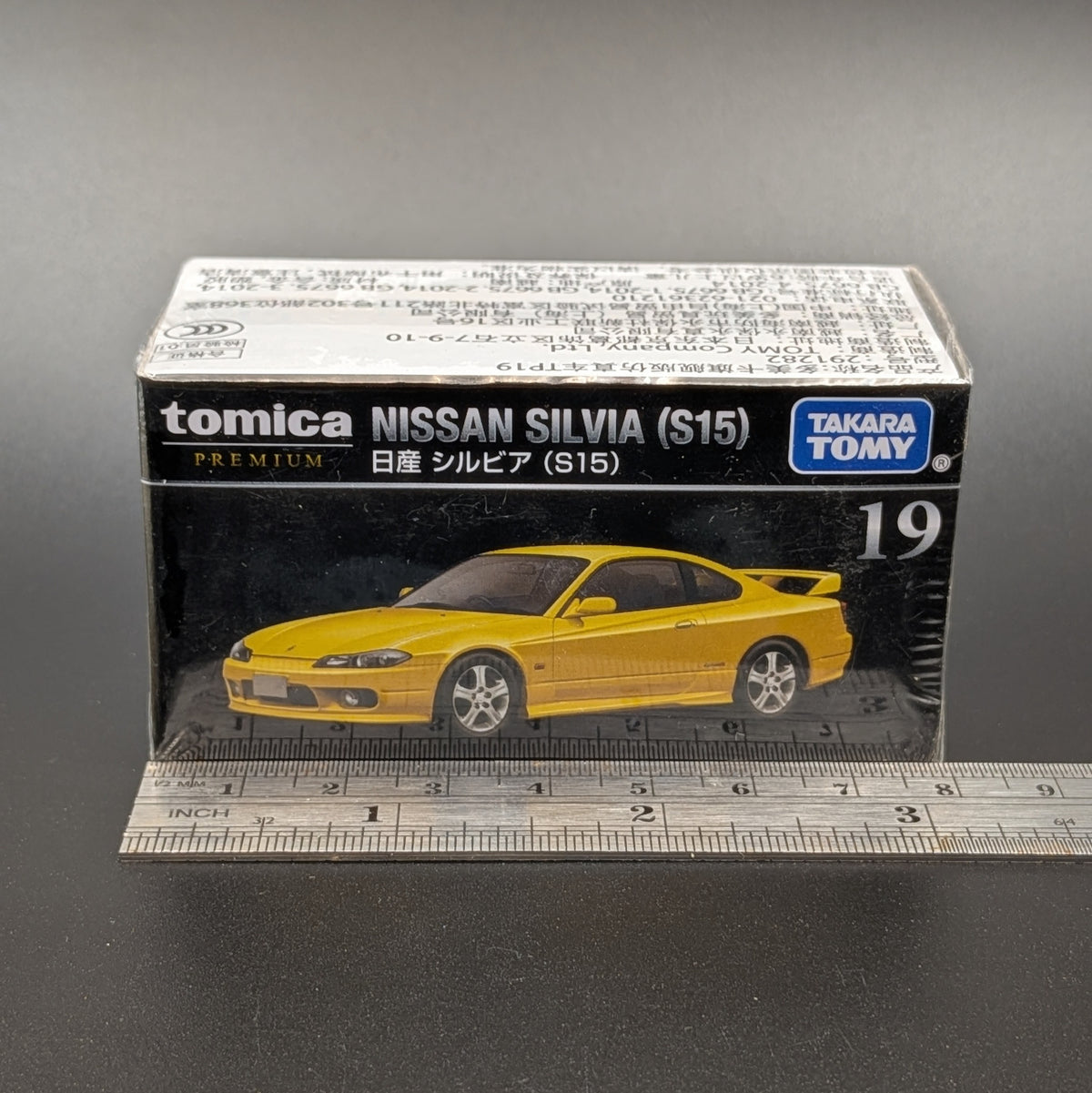 A boxed Tomica Premium Nissan Silvia (S15) model car, number 19, in its original packaging with a ruler at the bottom for scale, on a dark gray background.