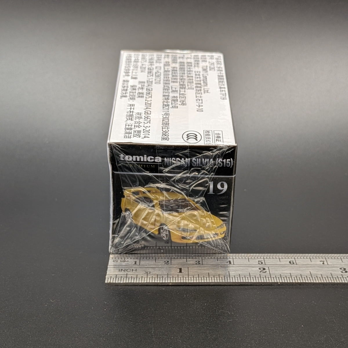 A boxed Tomica toy car wrapped in clear plastic, standing on a dark surface with a metal ruler along the bottom for scale. The box is mostly white and black with a small yellow car illustration on the front.