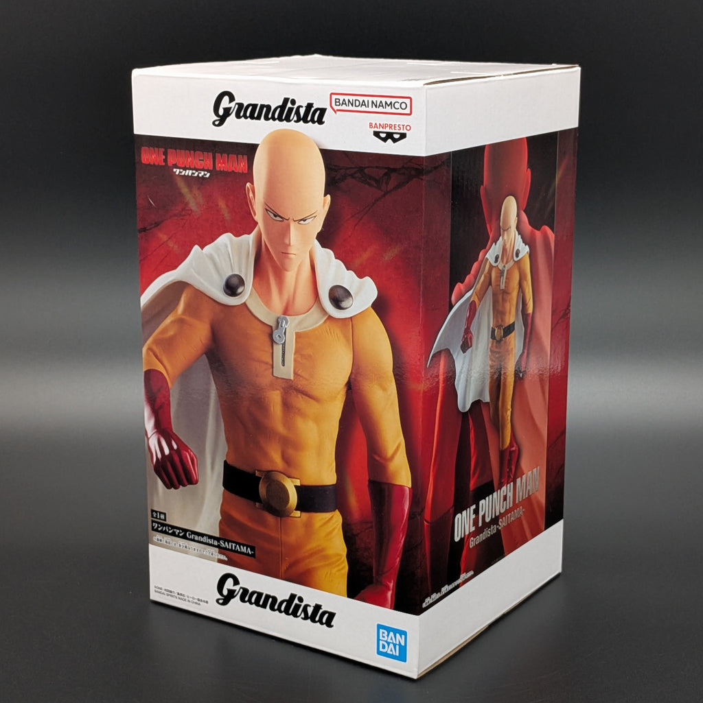 Bandai Namco Grandista One Punch Man Saitama 10-inch collectible figure in box, featuring Saitama in his signature yellow and red outfit with a white cape, set against a dark gray background.