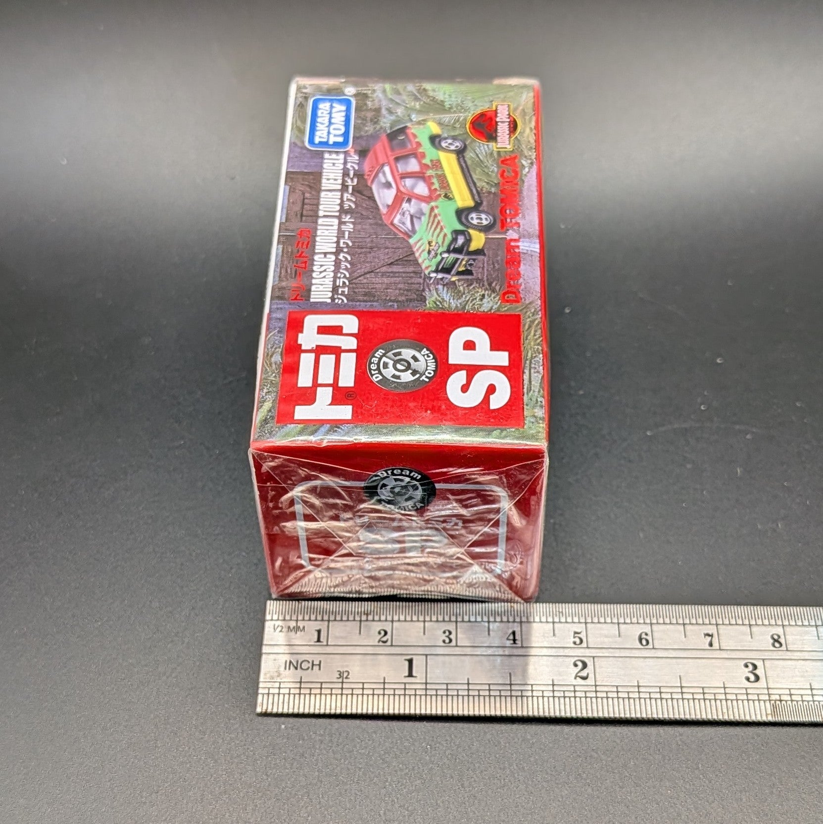 
Takara Tomy Dream Tomica SP Jurassic World Tour Vehicle (Ford Explorer), 1/64 scale diecast boxed and unopened. The picture is taken from slightly above and in front of the narrow side of the box, the metal ruler beside it shows a width of 4cm. Displayed on a black background