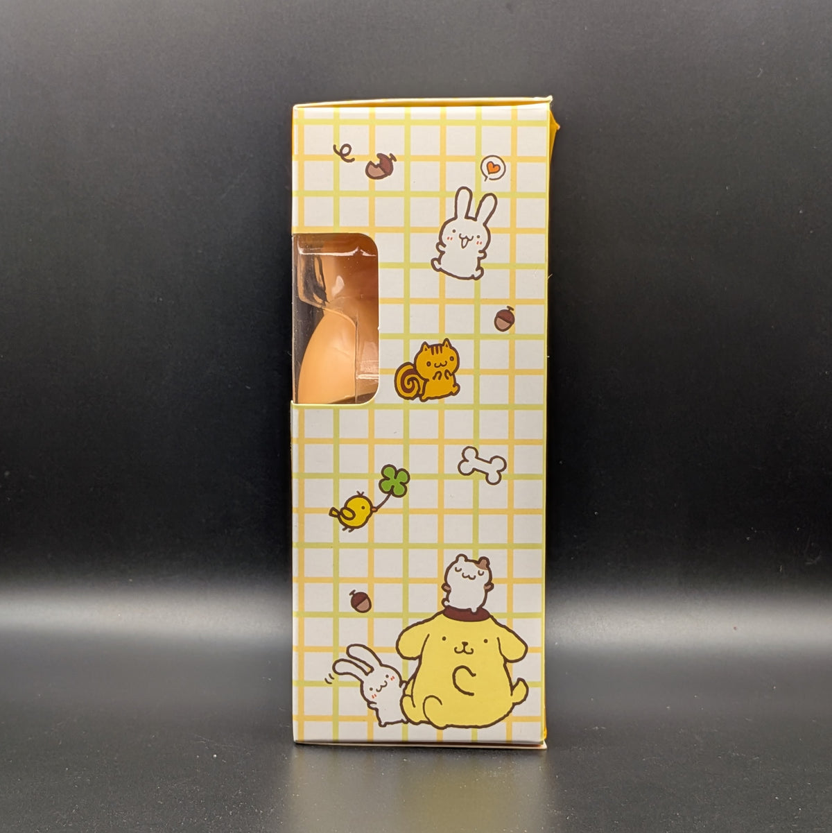 A tall rectangular product box with a yellow grid pattern and cute cartoon illustrations, including a rabbit, cat, and dog, standing upright against a dark gray background. A clear window on the left side reveals a small orange item inside.