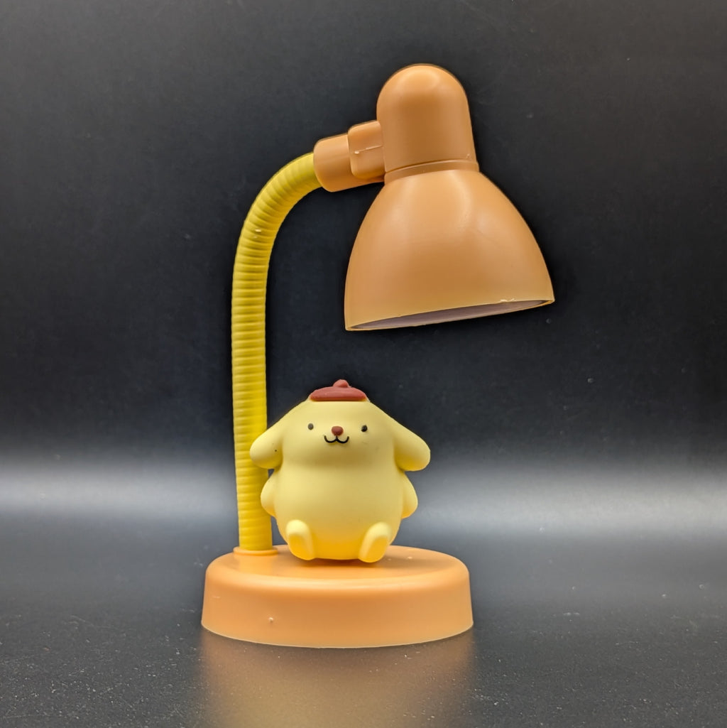 A small yellow character figurine sitting on an orange table lamp base, with the lamp's flexible yellow arm and orange shade above it, photographed against a dark gray background.