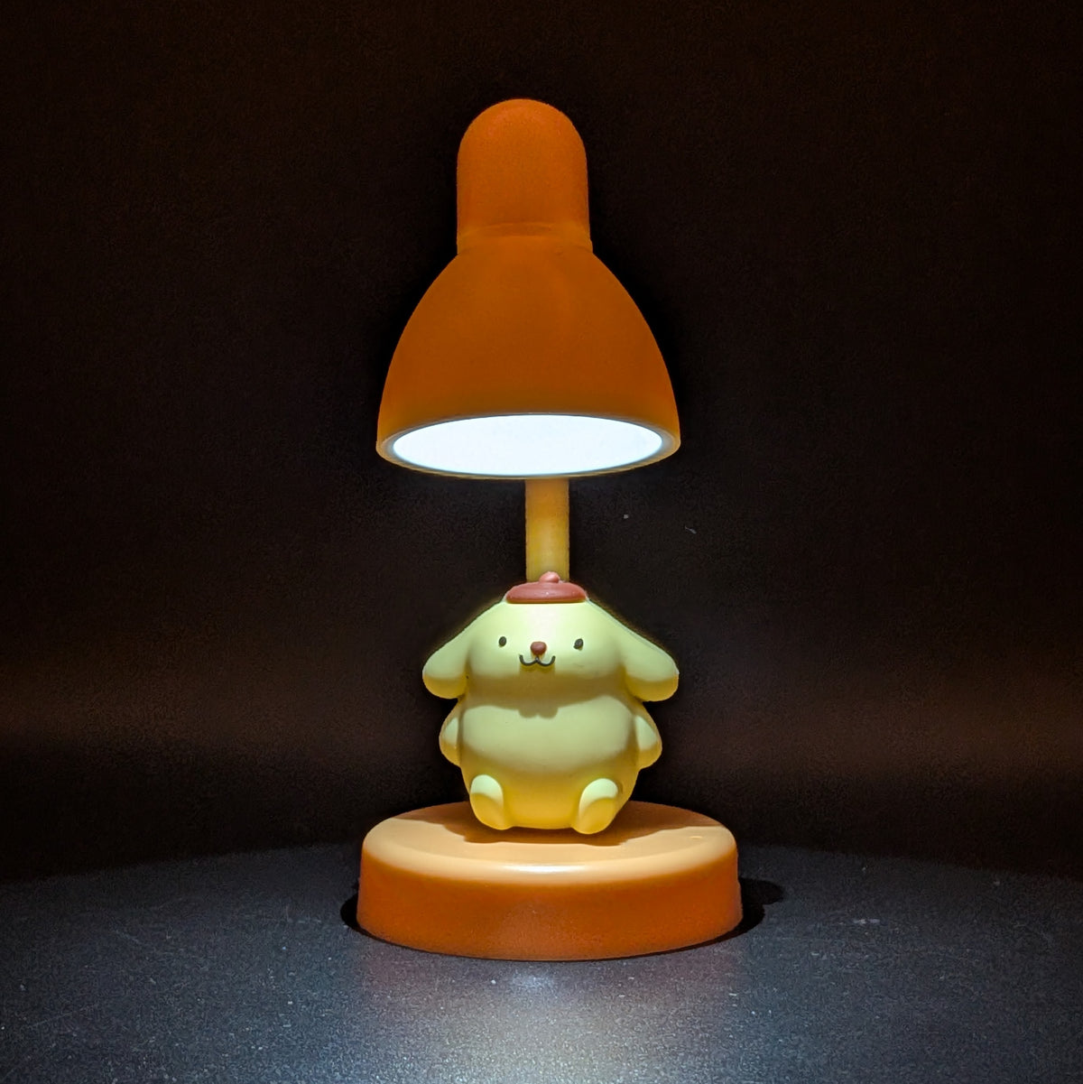 A cute cartoon table lamp featuring a yellow character sitting on an orange base, with an orange lampshade that glows with a white light, set against a dark background.