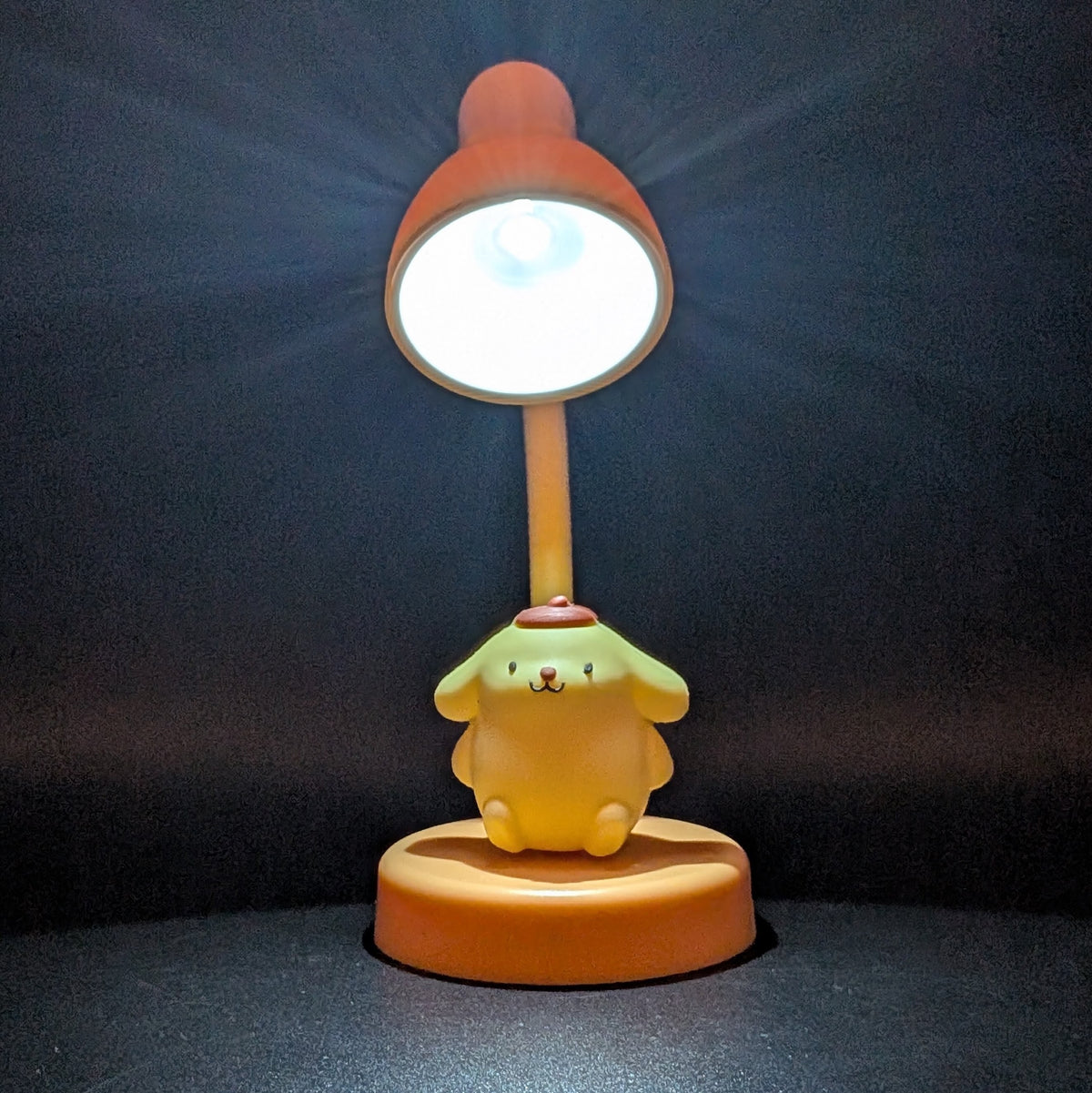 A cute desk lamp featuring a yellow cartoon dog character sitting on an orange circular base, with a flexible stem leading to an orange lamp head that is turned on and casting a bright glow.