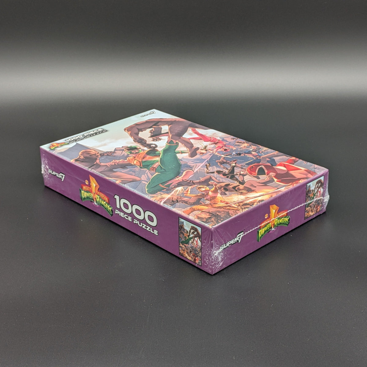 A sealed jigsaw puzzle box on a dark gray studio background. The box is purple with colorful fantasy artwork and large white text reading 1000 PIECE PUZZLE.