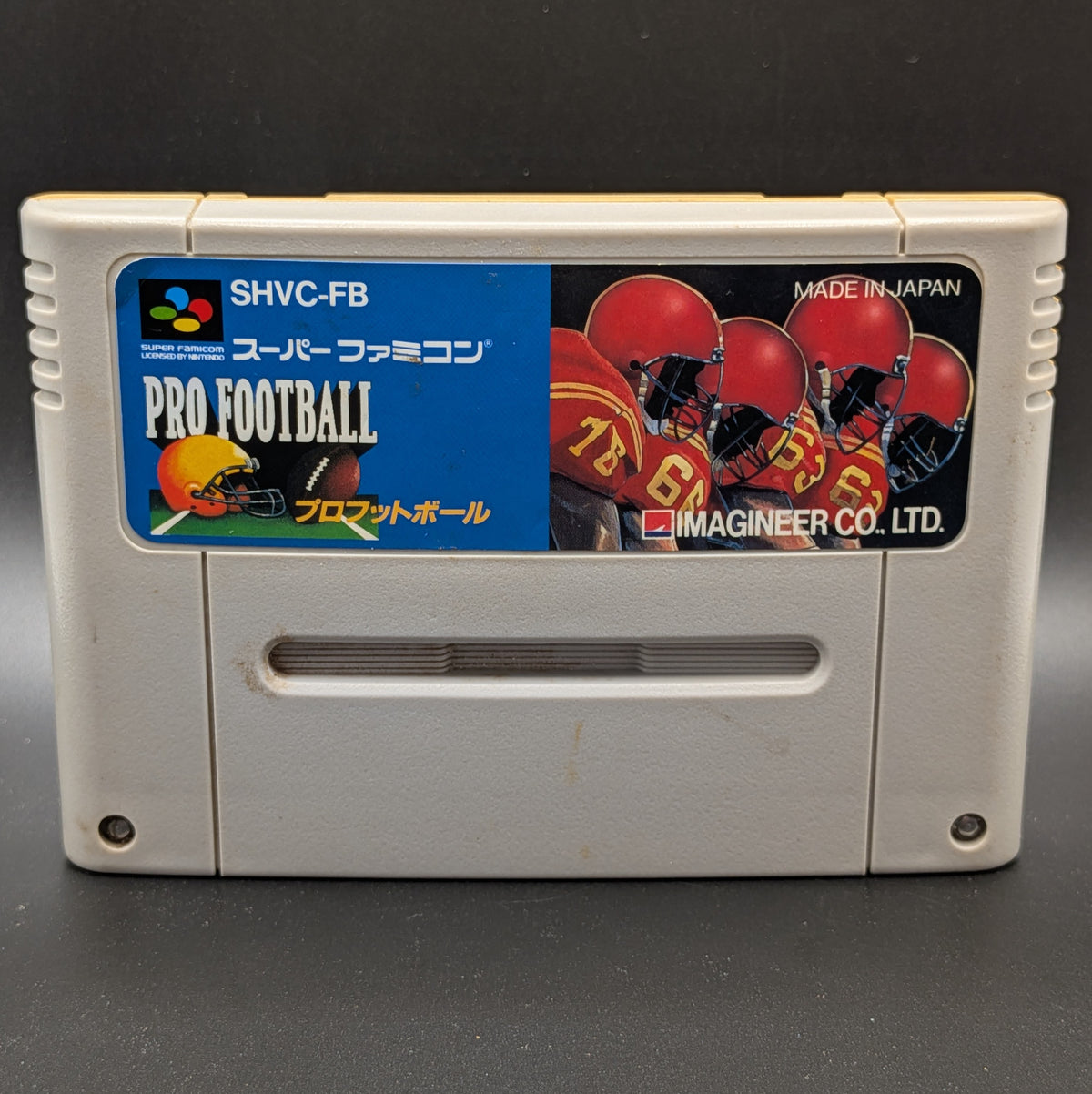 A close up of Pro Football (SHVC-FB) video game cartridge for the Super Famicom Entertainment System, shown standing up and facing forward on a dark grey background.
