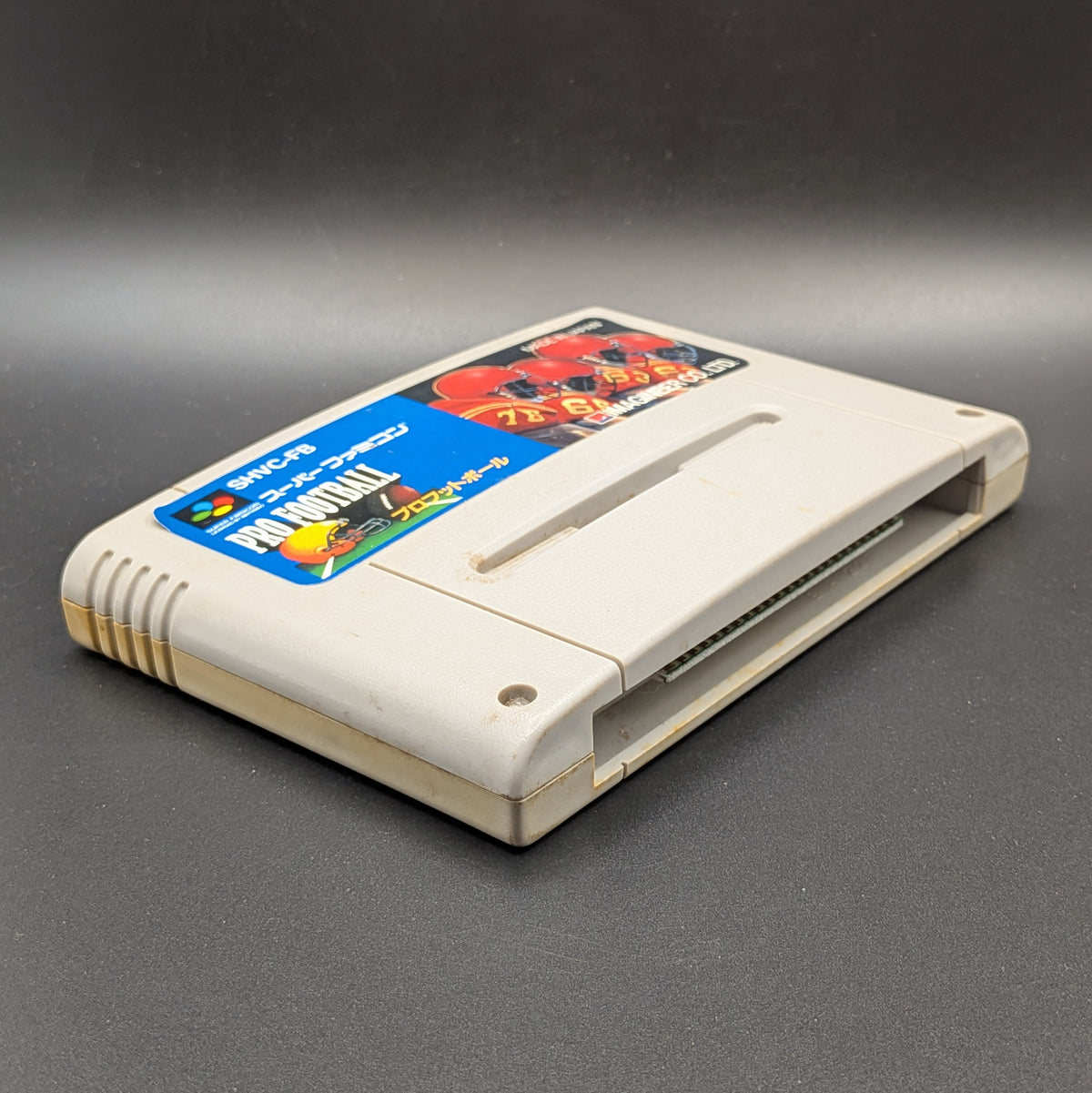 Pro Football (SHVC-FB) video game cartridge for the Super Famicom Entertainment System, shown laying on its back with the label facing up, at a 3/4 angle. Pictured on a dark grey background.