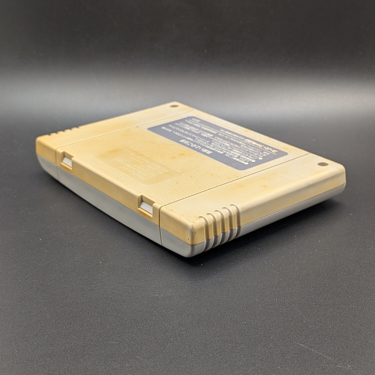 Pro Football (SHVC-FB) video game cartridge for the Super Famicom Entertainment System, shown laying on its front, at a 3/4 angle facing down so the back label is visible. Pictured on a dark grey background.