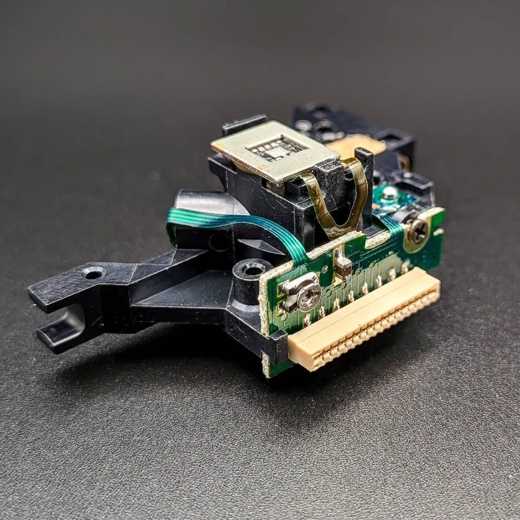 A close-up of a small electronic component or connector module with a black plastic housing, green circuit board, gold metal contacts, and a beige ribbon connector, photographed on a dark textured surface.