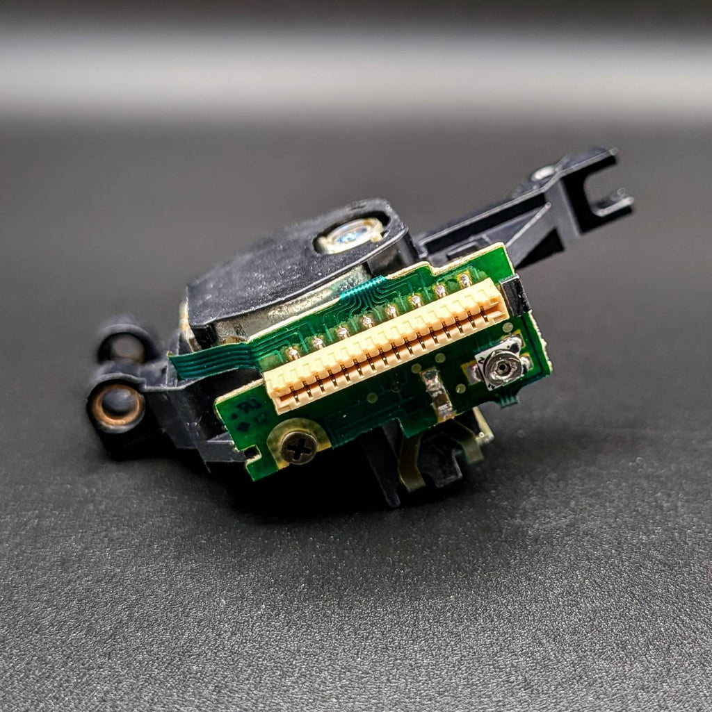 A close-up of a small electronic component or connector assembly with a green circuit board, gold-plated pins, black plastic housing, and a small silver screw, photographed on a dark gray surface.