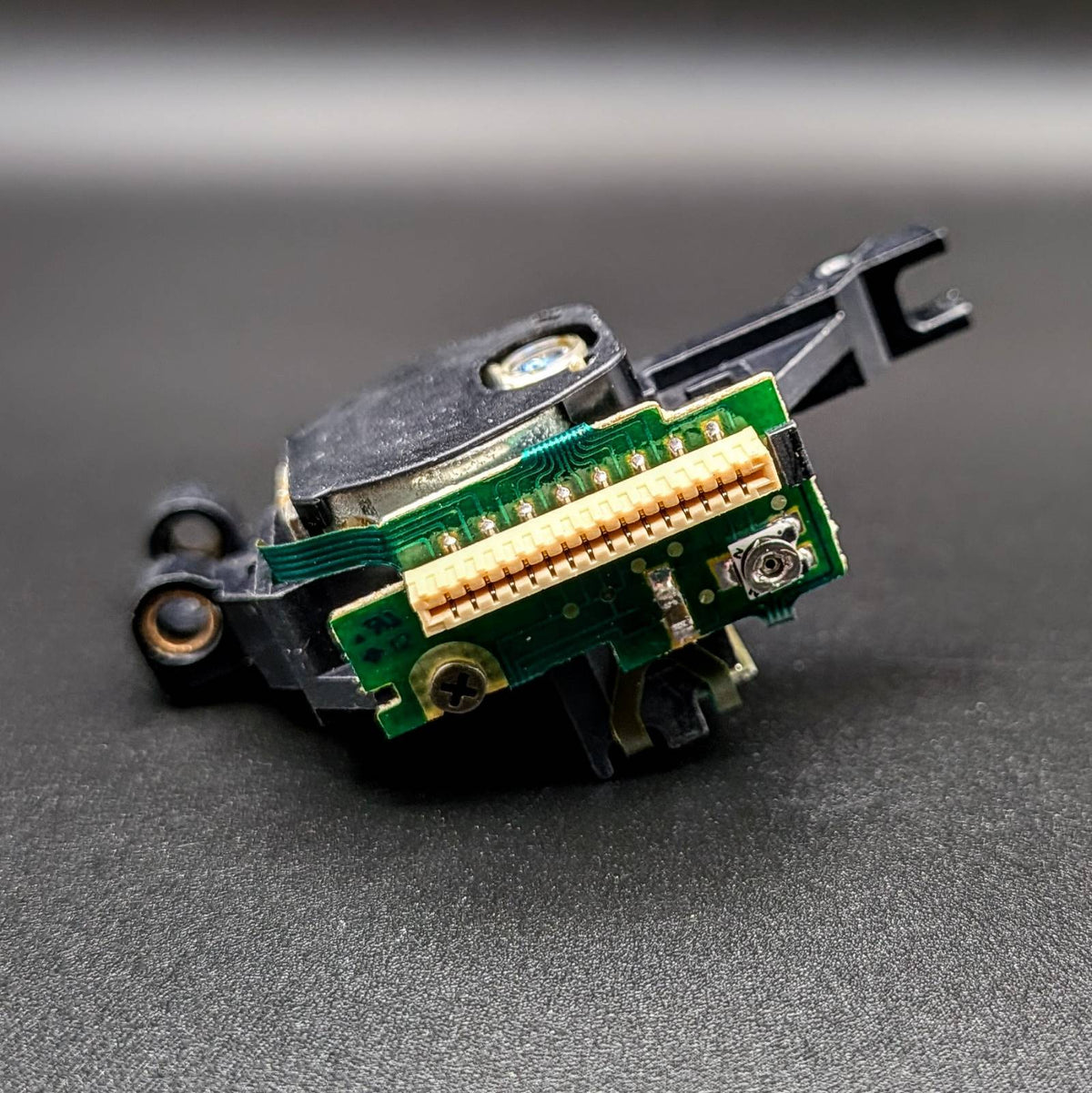 A close-up of a small electronic component or connector assembly with a green circuit board, gold-plated pins, black plastic housing, and a small silver screw, photographed on a dark gray surface.