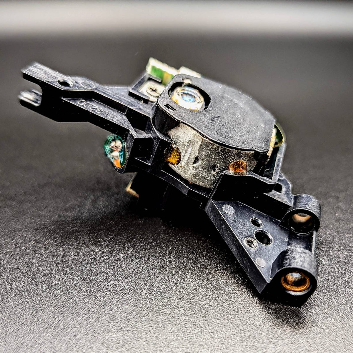 A close-up of a black plastic optical laser with small green circuitry, and metallic details, resting on a dark textured surface.