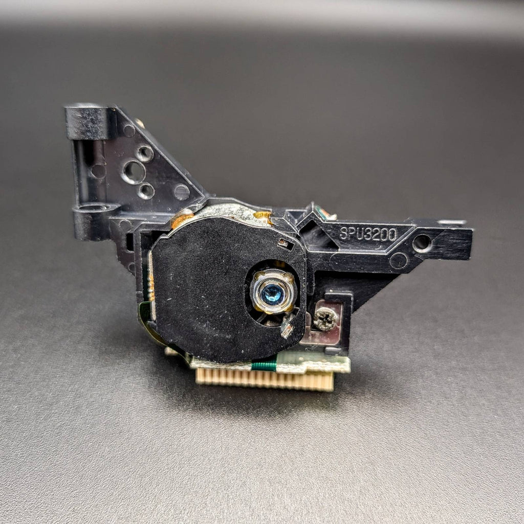 A replacement electronic component, specifically a black plastic laser  lens housing with a gold PCI-E style connector and a blue-coated lens, shown on a dark gray background.
