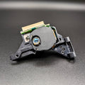 A close-up of a black plastic SEGA Dreamcast optical assembly with a gold connector and a blue lens, photographed on a dark gray textured surface.
