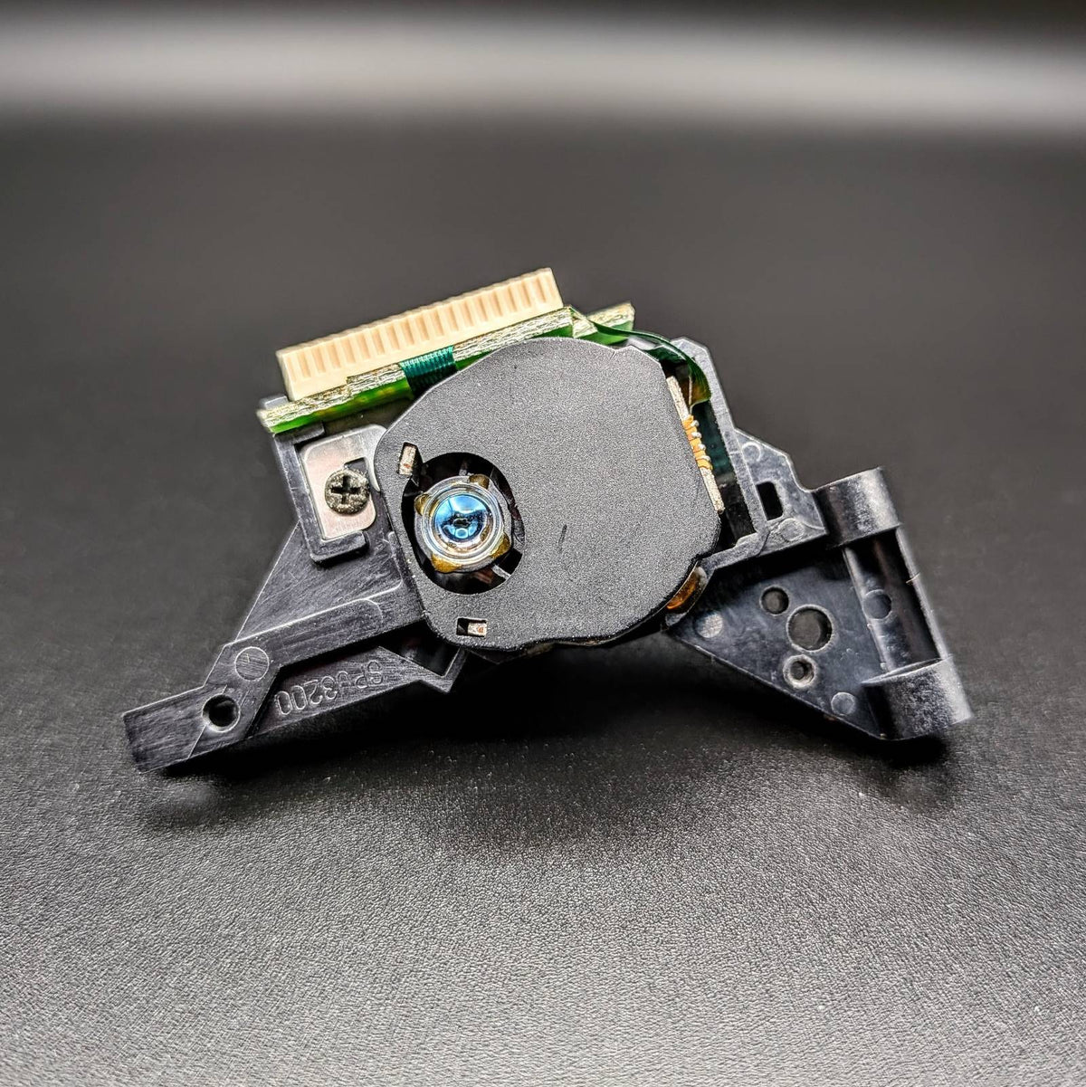 A close-up of a black plastic SEGA Dreamcast optical assembly with a gold connector and a blue lens, photographed on a dark gray textured surface.