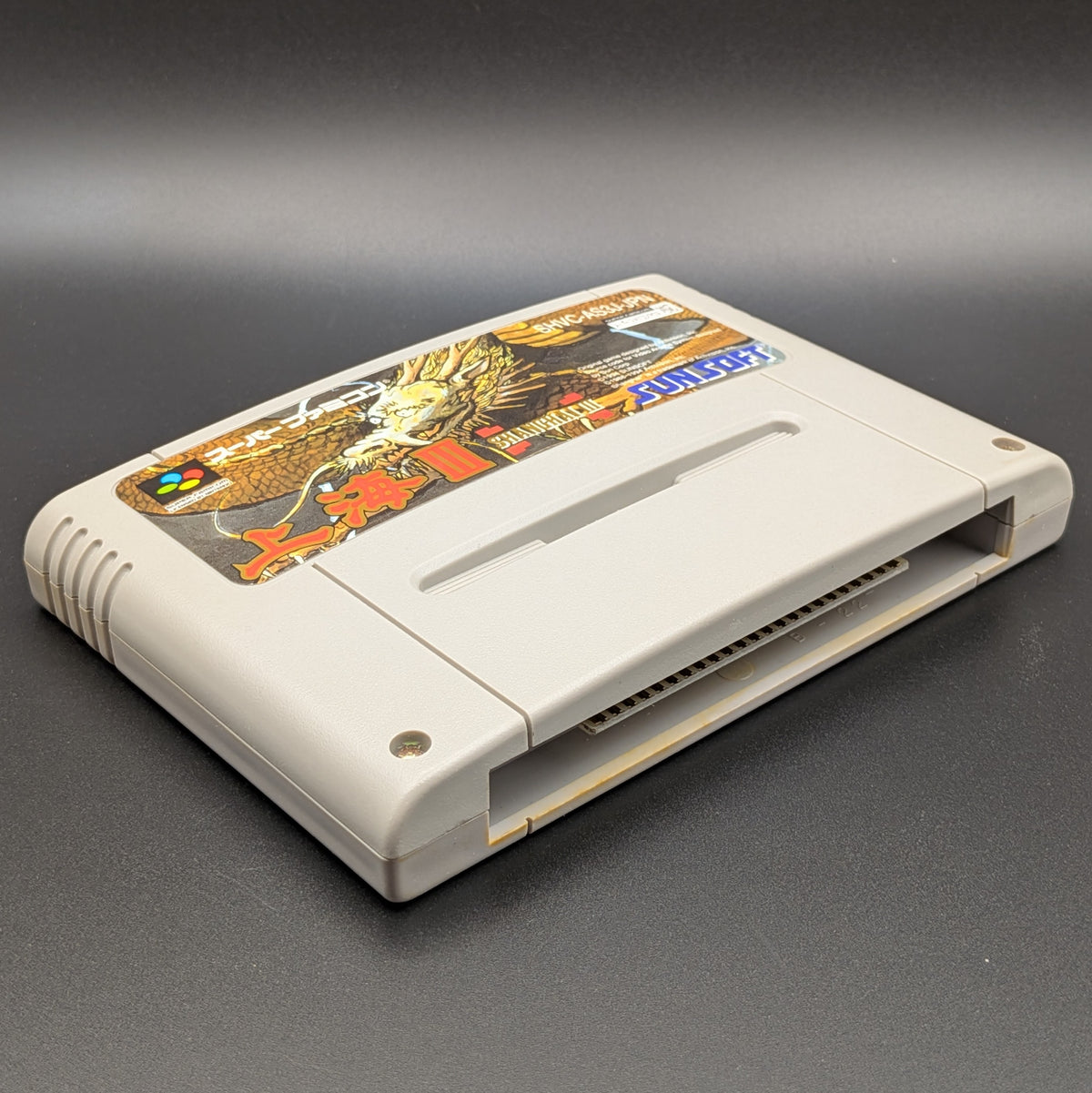 Shanghai 3 video game cartridge for Nintendo's Super Famicom (SFC) Entertainment System, shown laying on its back with the label facing upwards. Showing the left hand side and bottom slot. It is displayed on a dark grey background.