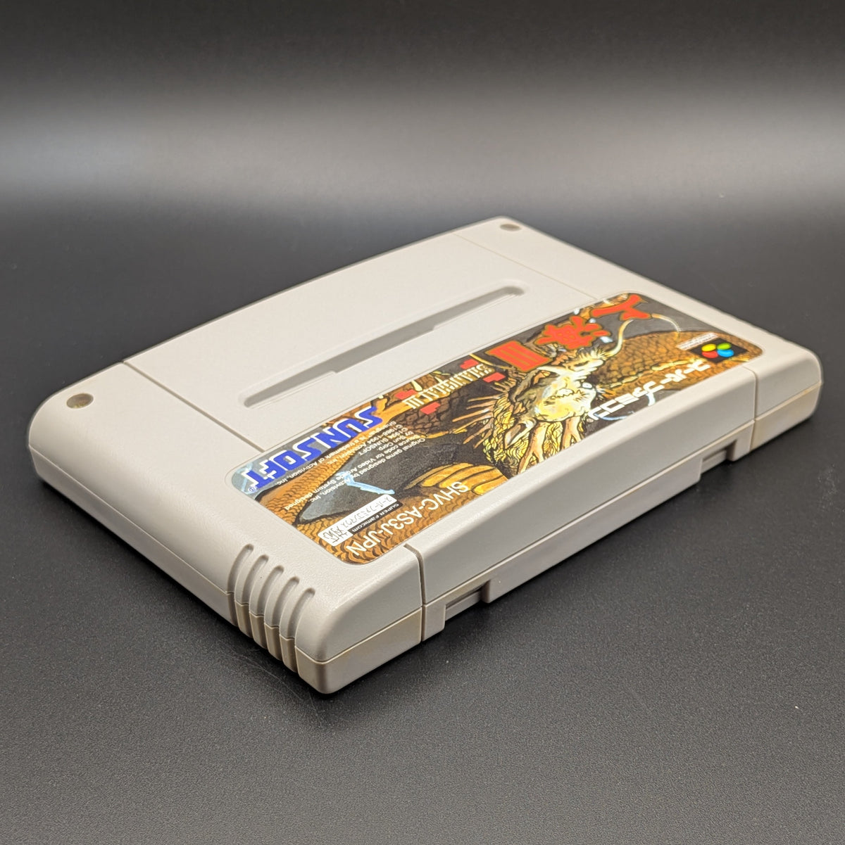  Shanghai 3 video game cartridge for Nintendo's Super Famicom (SFC) Entertainment System, shown laying on its back with the label facing upwards. Showing the right hand side and top edge. It is displayed on a dark grey background.