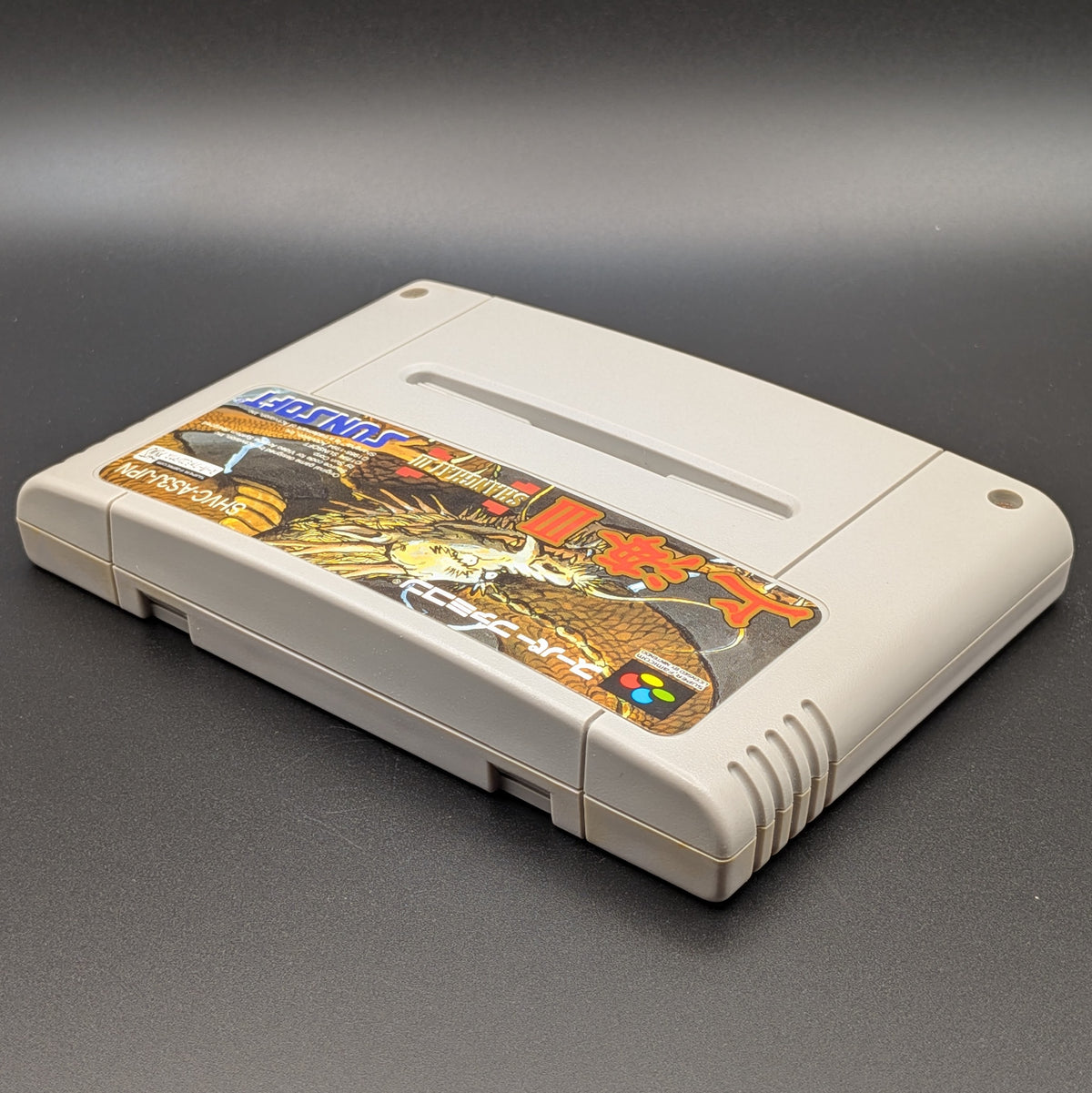Shanghai 3 video game cartridge for Nintendo's Super Famicom (SFC) Entertainment System, shown laying on its back with the label facing upwards. Showing the left hand side and top edge. It is displayed on a dark grey background.